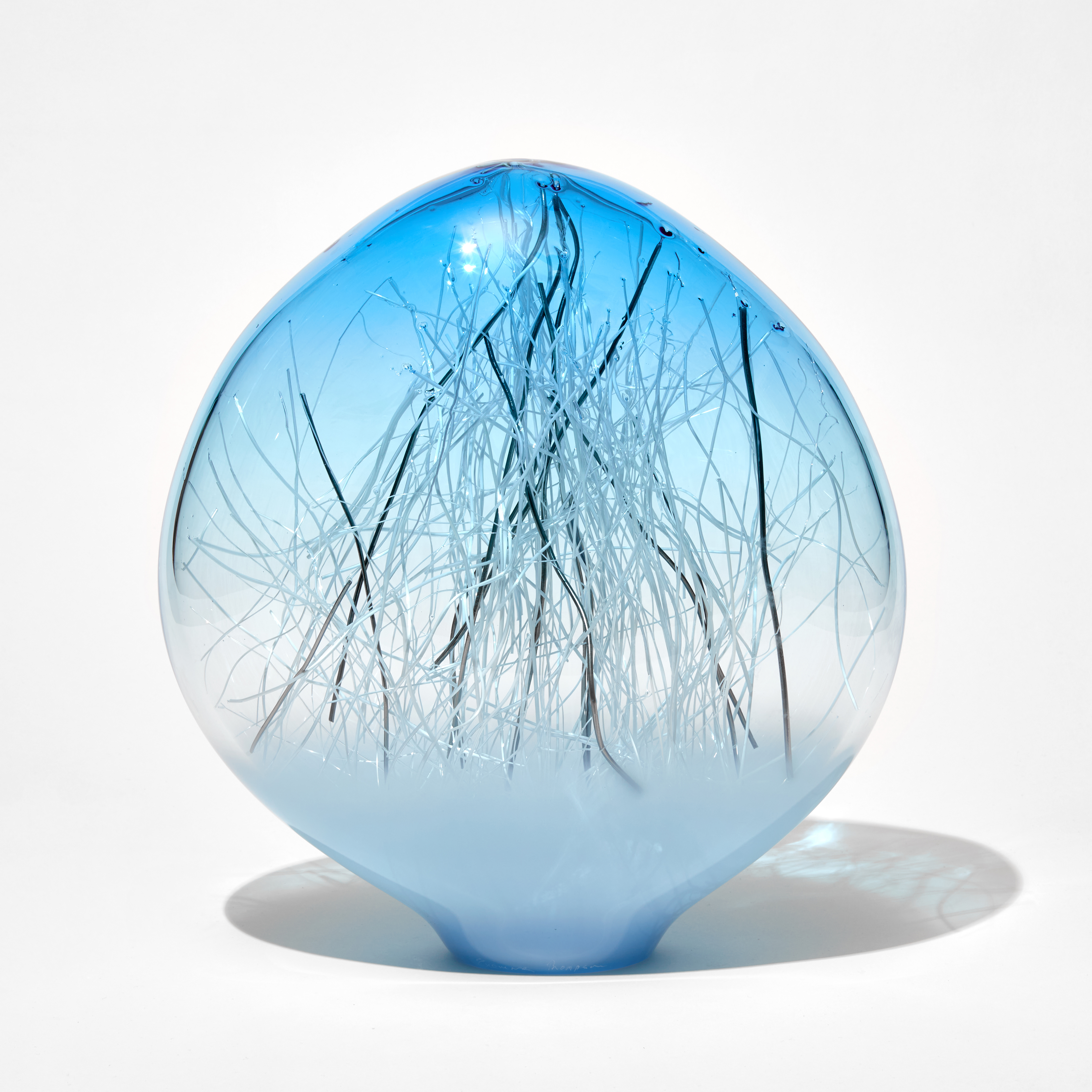 low ovoid hand made glass sculpture with soft blue base fading to clear to a bright aqua top with thin canes in white and platinum trapped inside