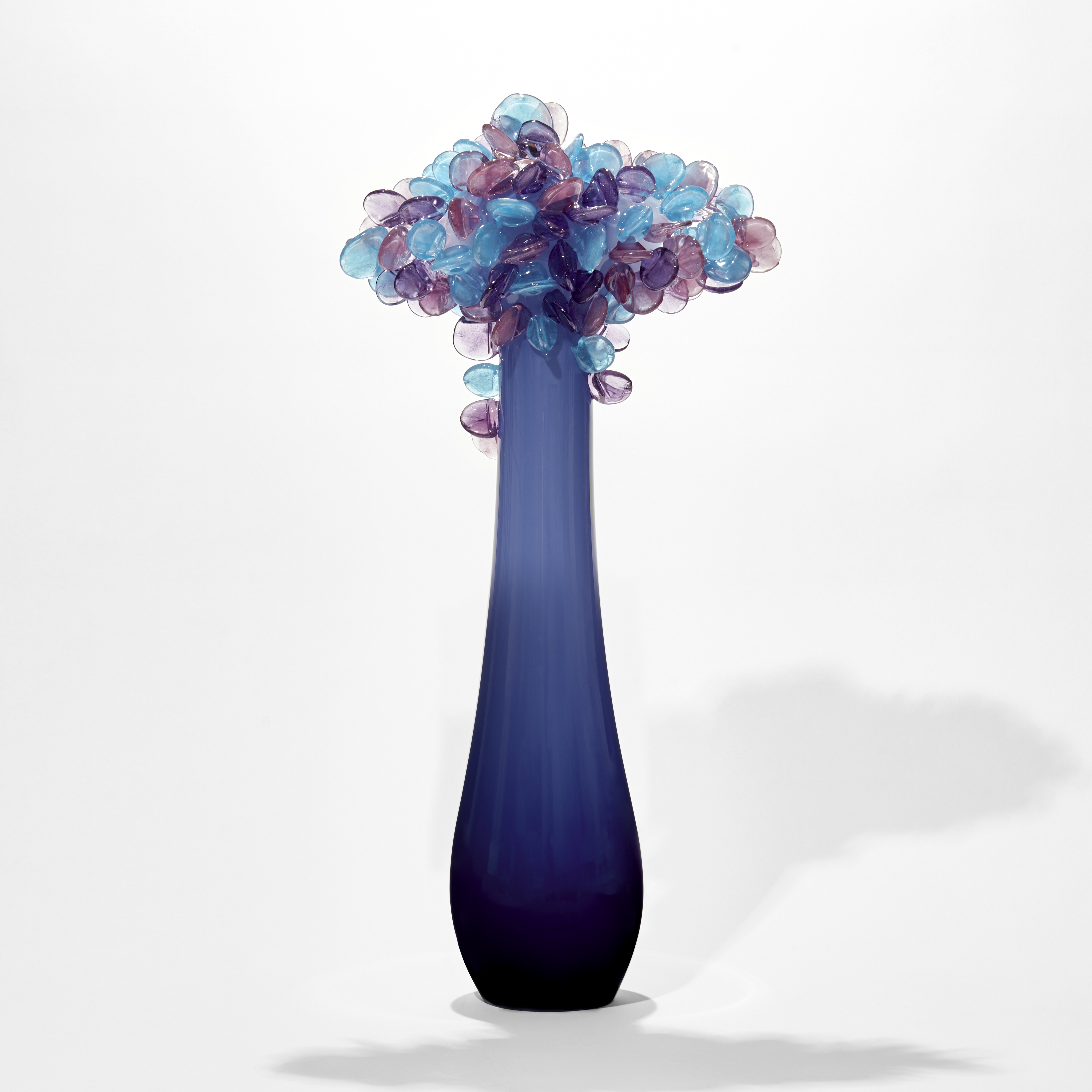 simplified tree with elongated tear shaped smooth trunk in dark blue lightening to the top with a cluster of lollipop leaves on top in blue and lilac handmade from glass