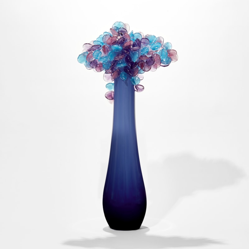 simplified tree with elongated tear shaped smooth trunk in dark blue lightening to the top with a cluster of lollipop leaves on top in blue and lilac handmade from glass