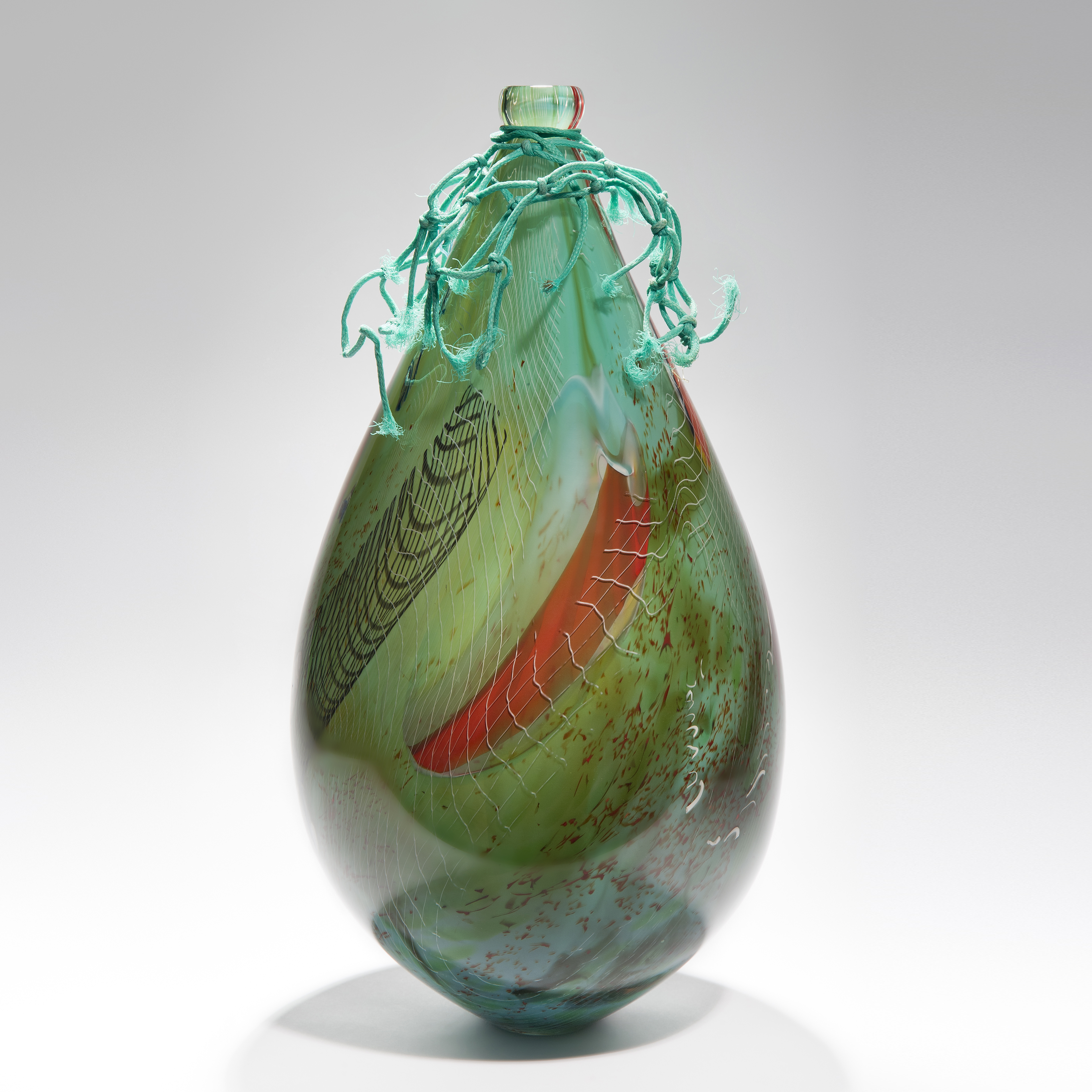 smooth droplet shaped green and reddish amber sculpture with lined areas of detail hand made from glass with ocean plastic cap and trim of found fishing net