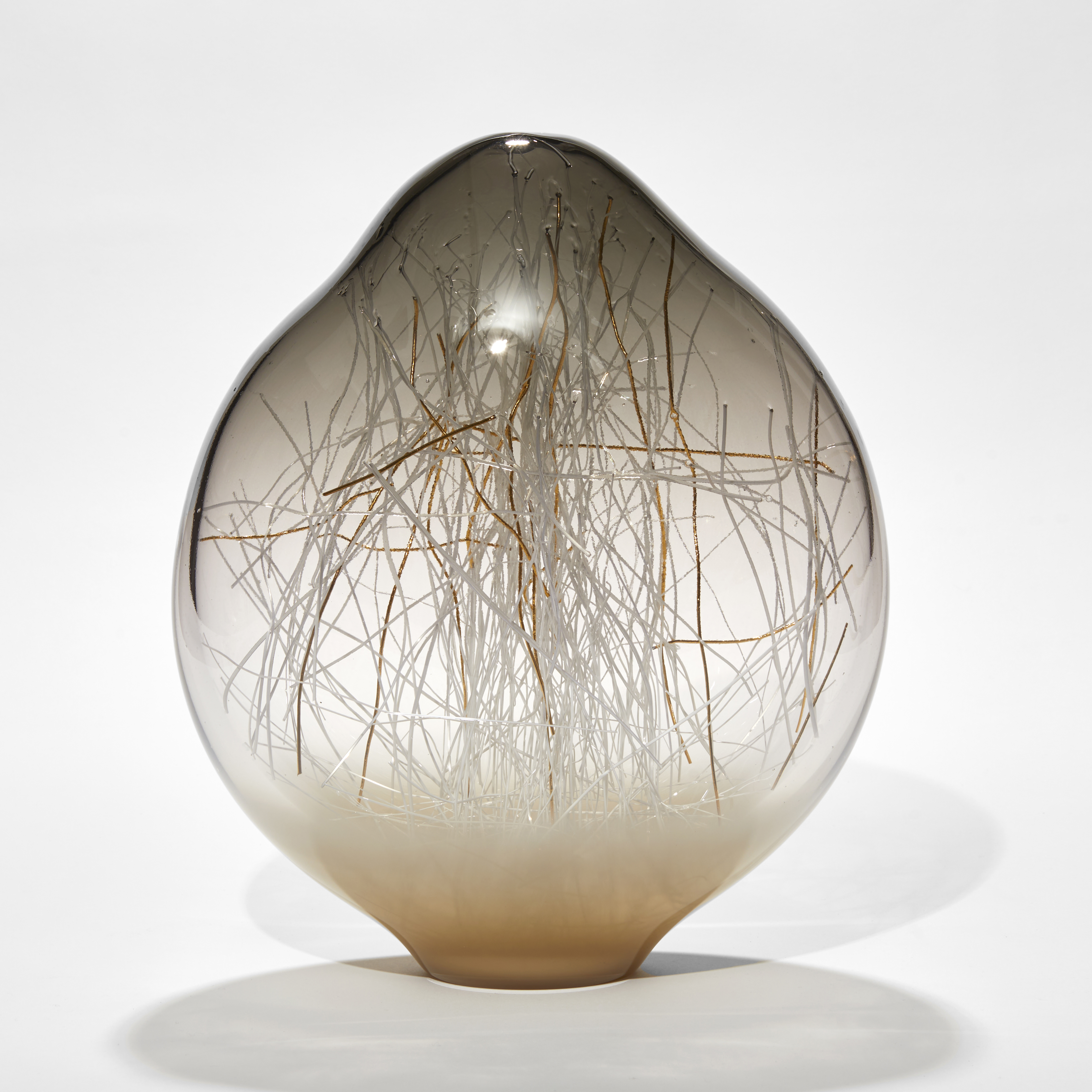 Amorphic shaped sculpture in transparent warm grey fading to clear then opaque taupe with organic canes in white and gold trapped inside hand made from glass