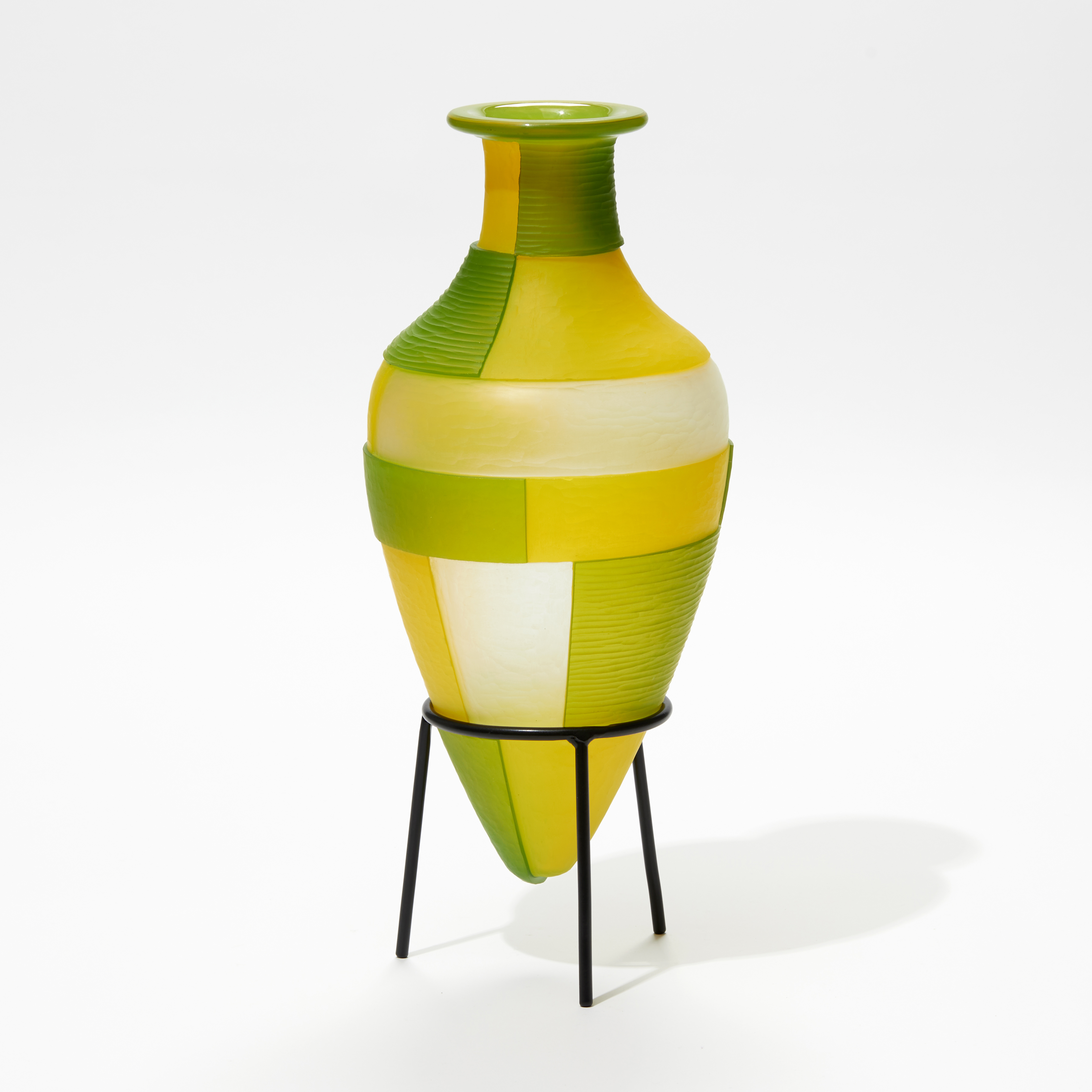 green and yellow amphora bottle with geometric square cut bold patterns on a black steel tripod stand handmade from glass