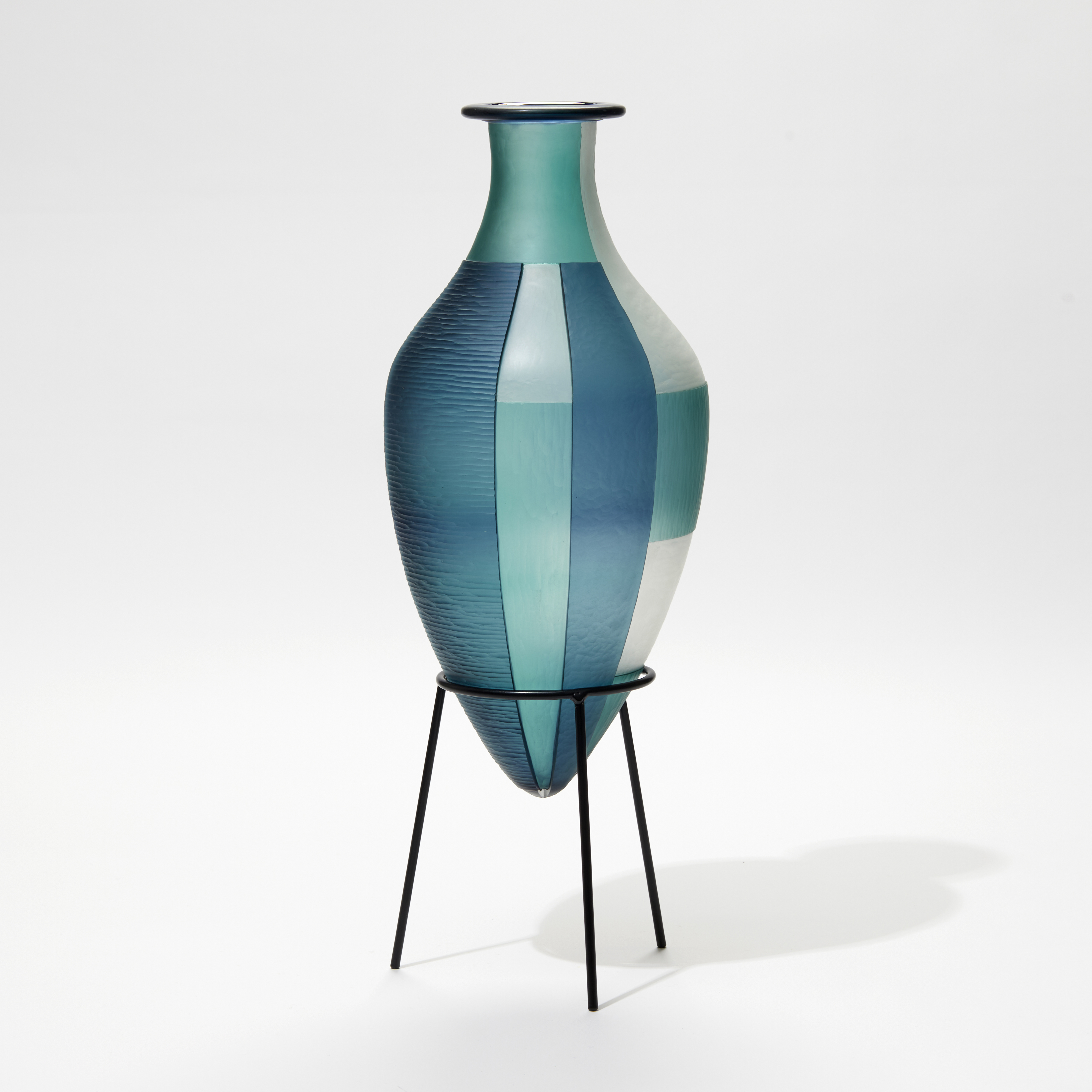 blue jade and white elongated bottled with pointed base and wide rim and an abstract block cut pattern hand made from glass with a black steel stand