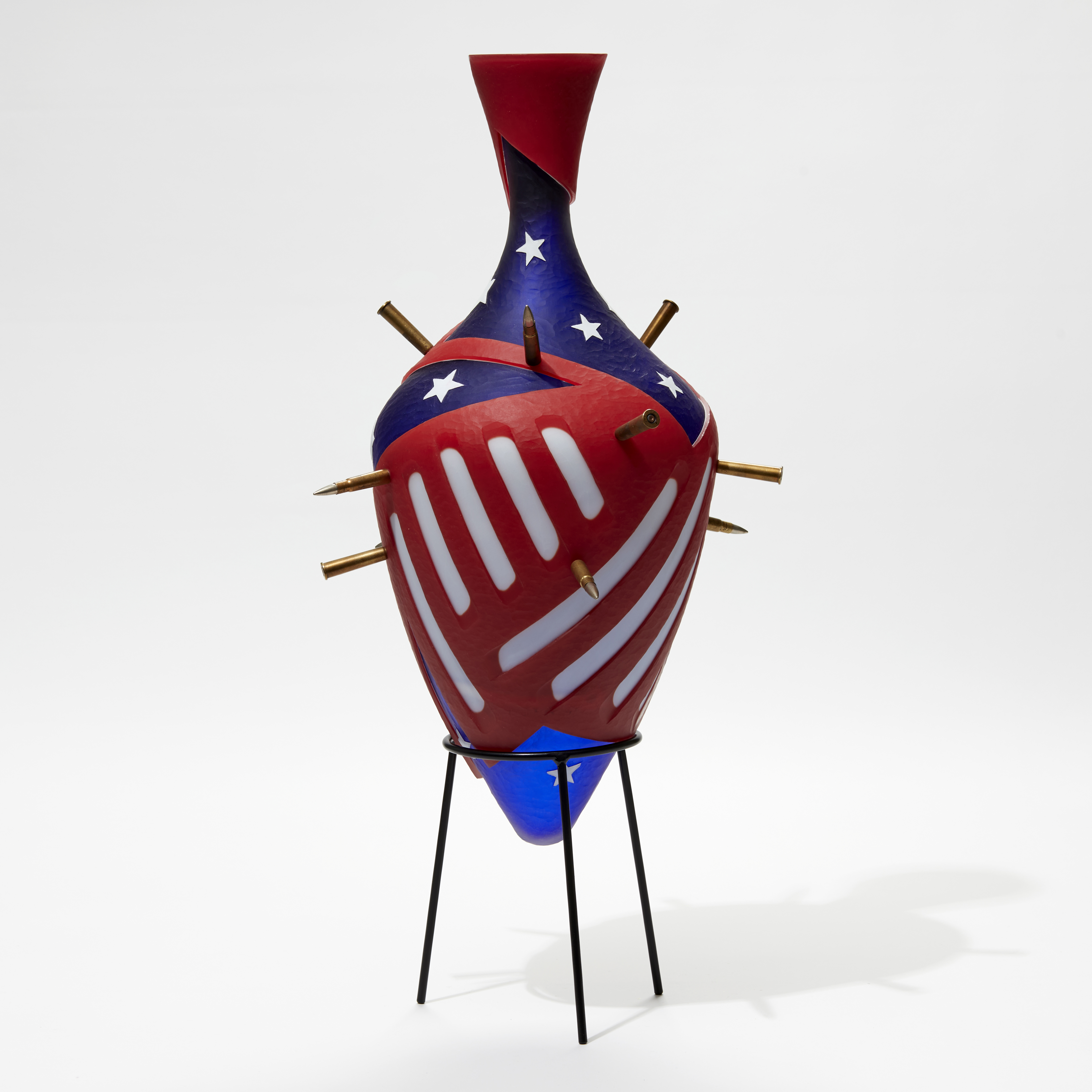 red white and blue amphora bottle with abstract stars and strips pattern pierced with bullets with a black metal stand