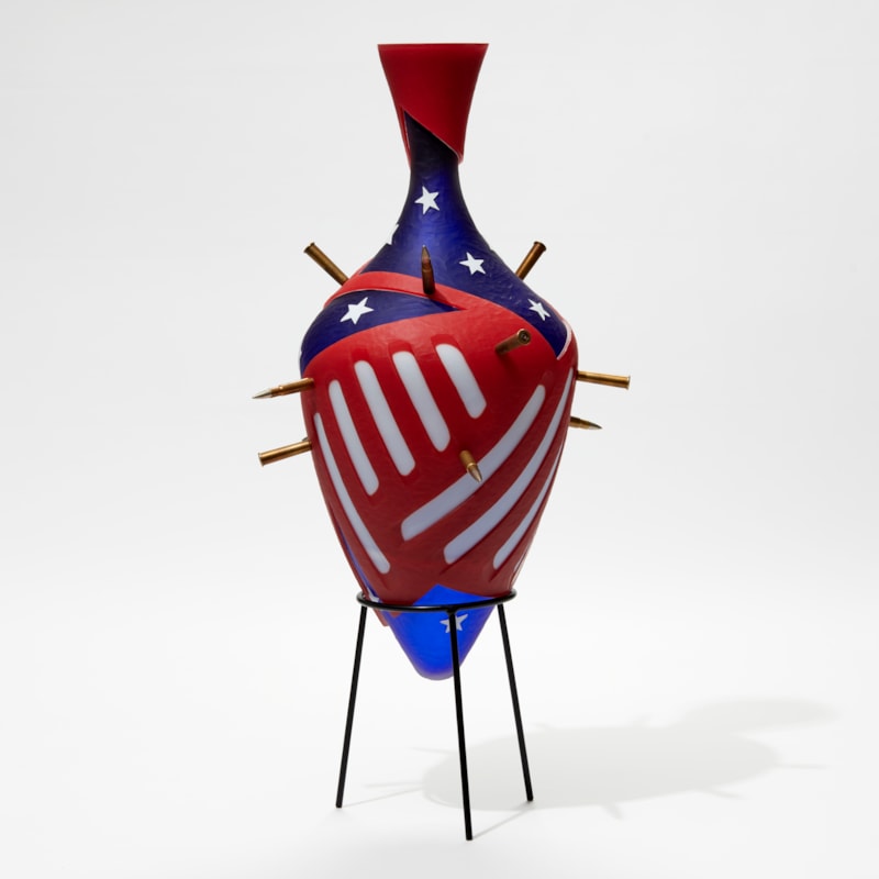 red white and blue amphora bottle with abstract stars and strips pattern pierced with bullets with a black metal stand