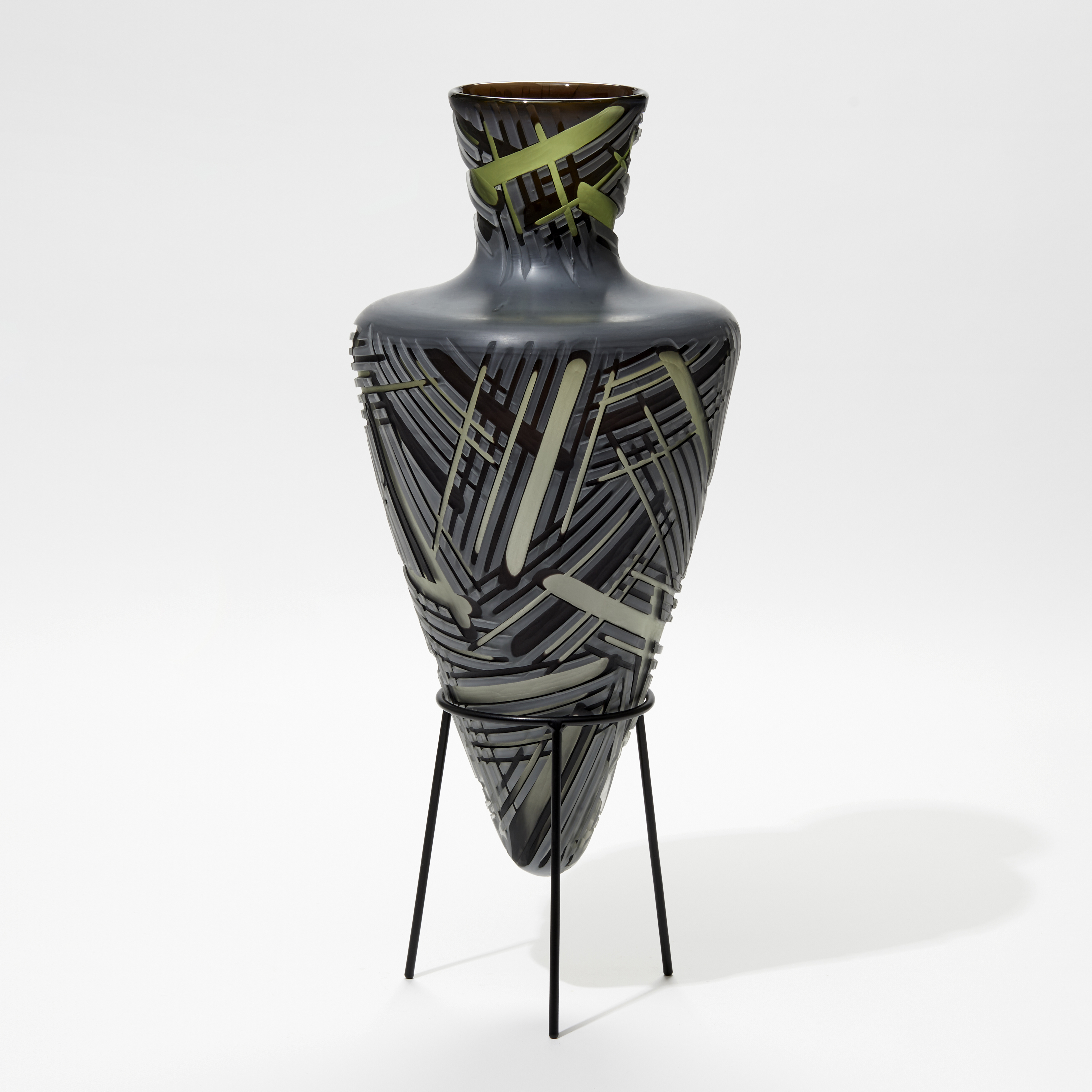 pointy bottomed bottle in dark grey and green which deep lines of differing widths cut in the surface hand made from glass with a steel stand 