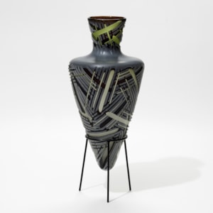 pointy bottomed bottle in dark grey and green which deep lines of differing widths cut in the surface hand made from glass with a steel stand