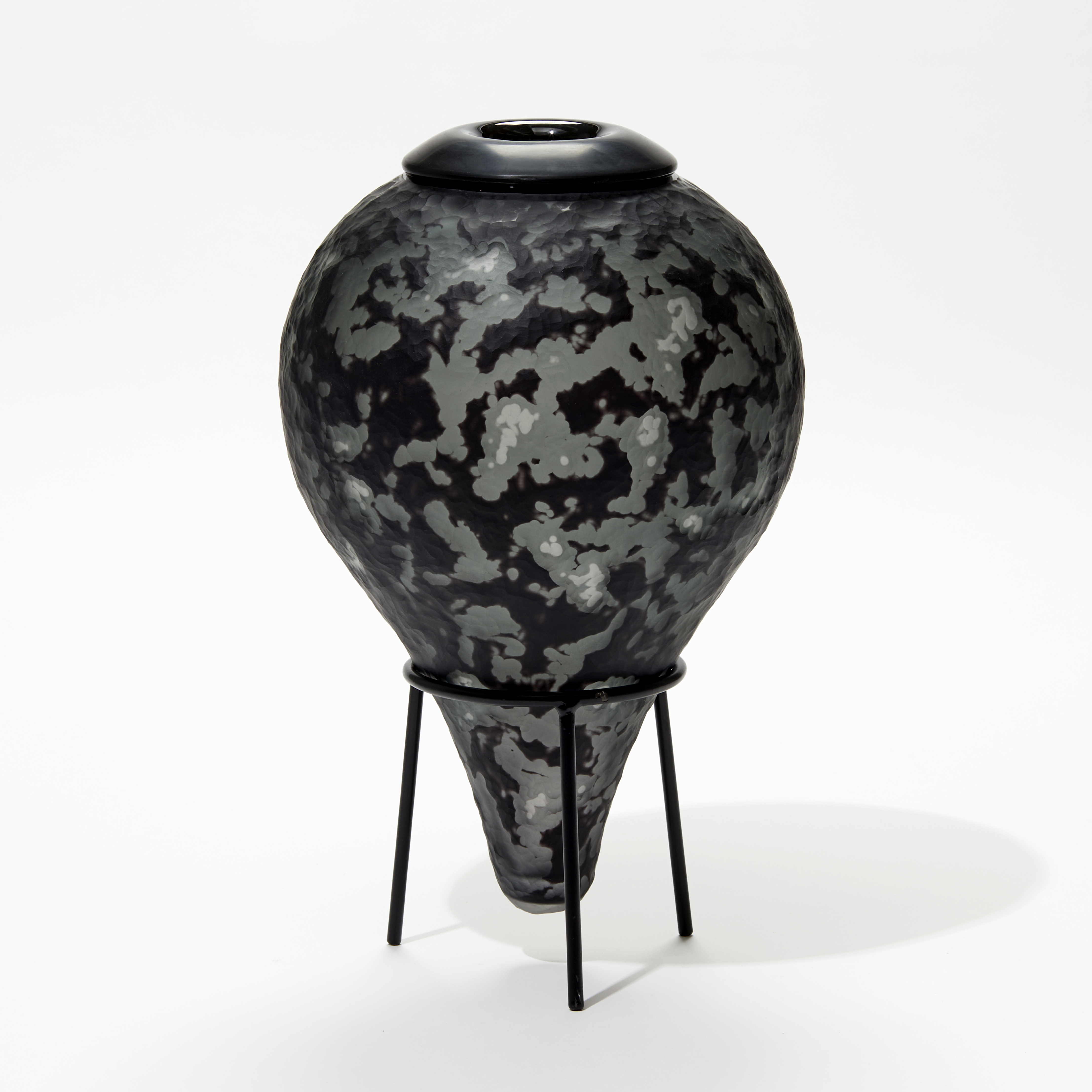 upside down teardrop shaped bottle in black and grey with textured surface and tripod black steel stand handmade from glass