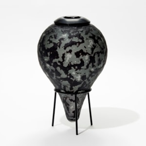 upside down teardrop shaped bottle in black and grey with textured surface and tripod black steel stand handmade from glass
