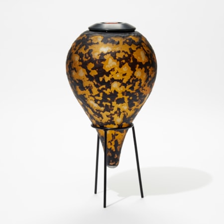 amber and black teardrop bottle with textured organic cut surface sat on a black tripod museum style stand
