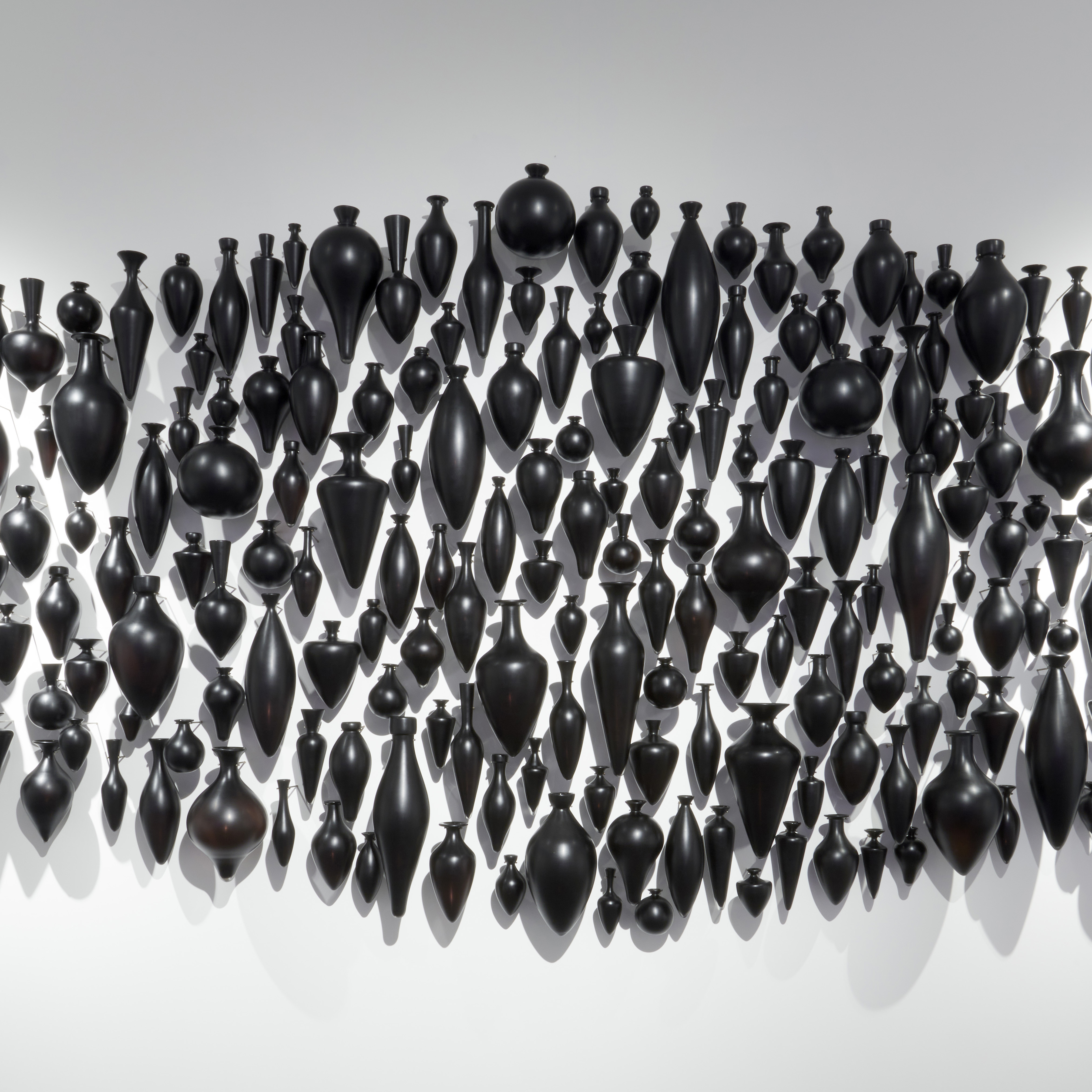 hundreds of black handblown and polished small amphora bottles wall mounted and hung in a clustered pointed horizontal oval configuration