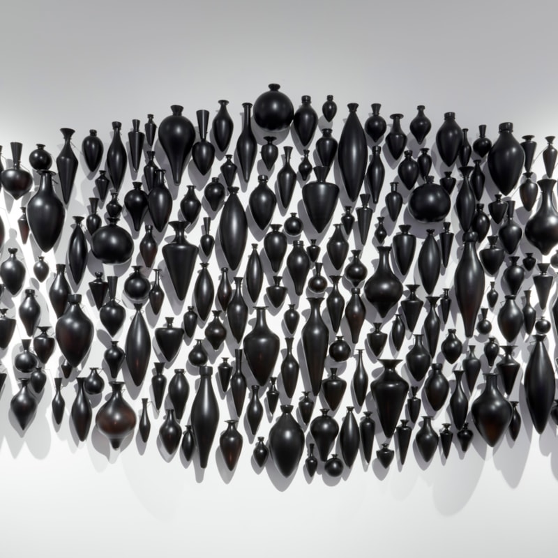 hundreds of black handblown and polished small amphora bottles wall mounted and hung in a clustered pointed horizontal oval configuration