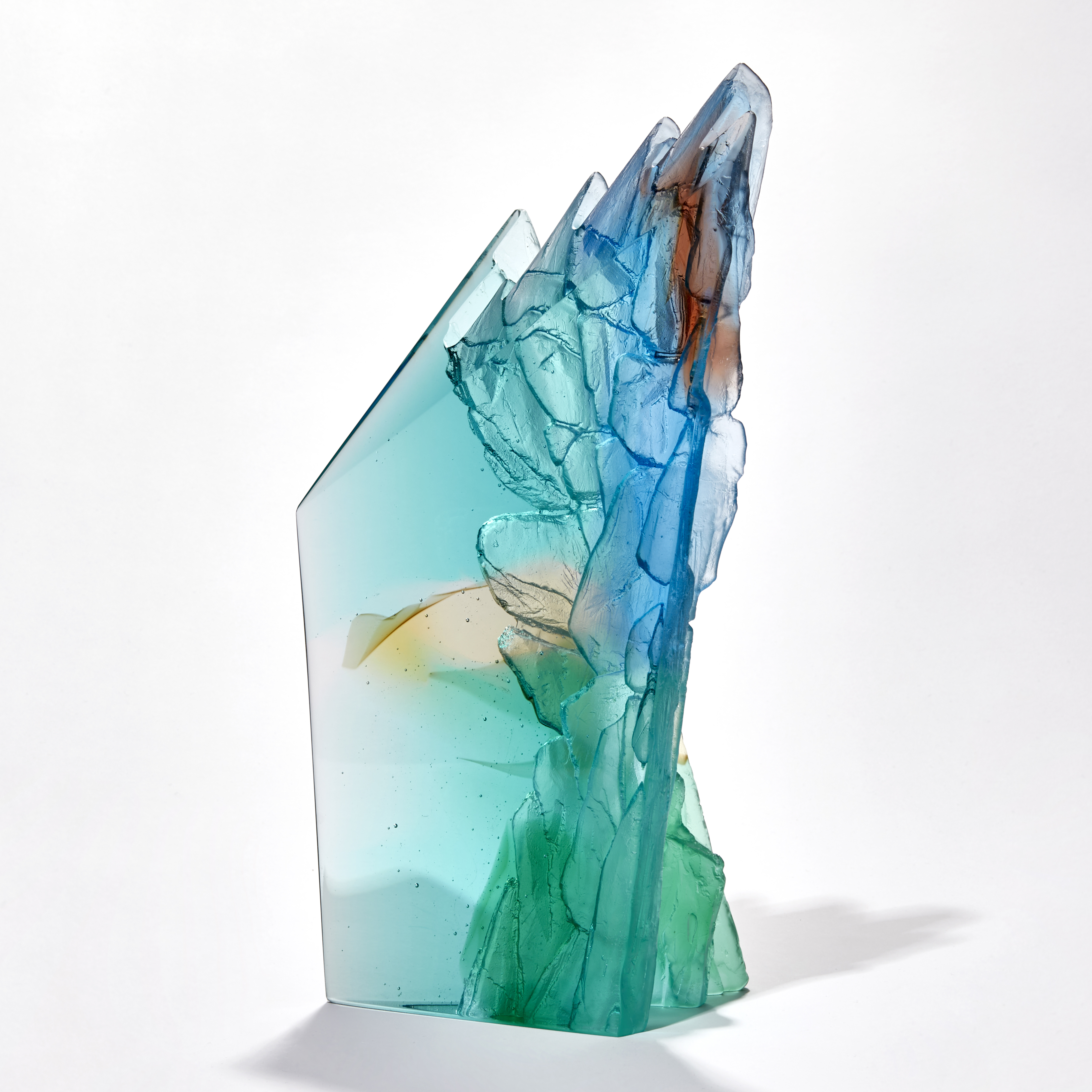 transparent abstract sculpture with two flat sheer sides and inner rugged cliff face textured edge in turquoise jade amber beige blue and red brown hand made from glass