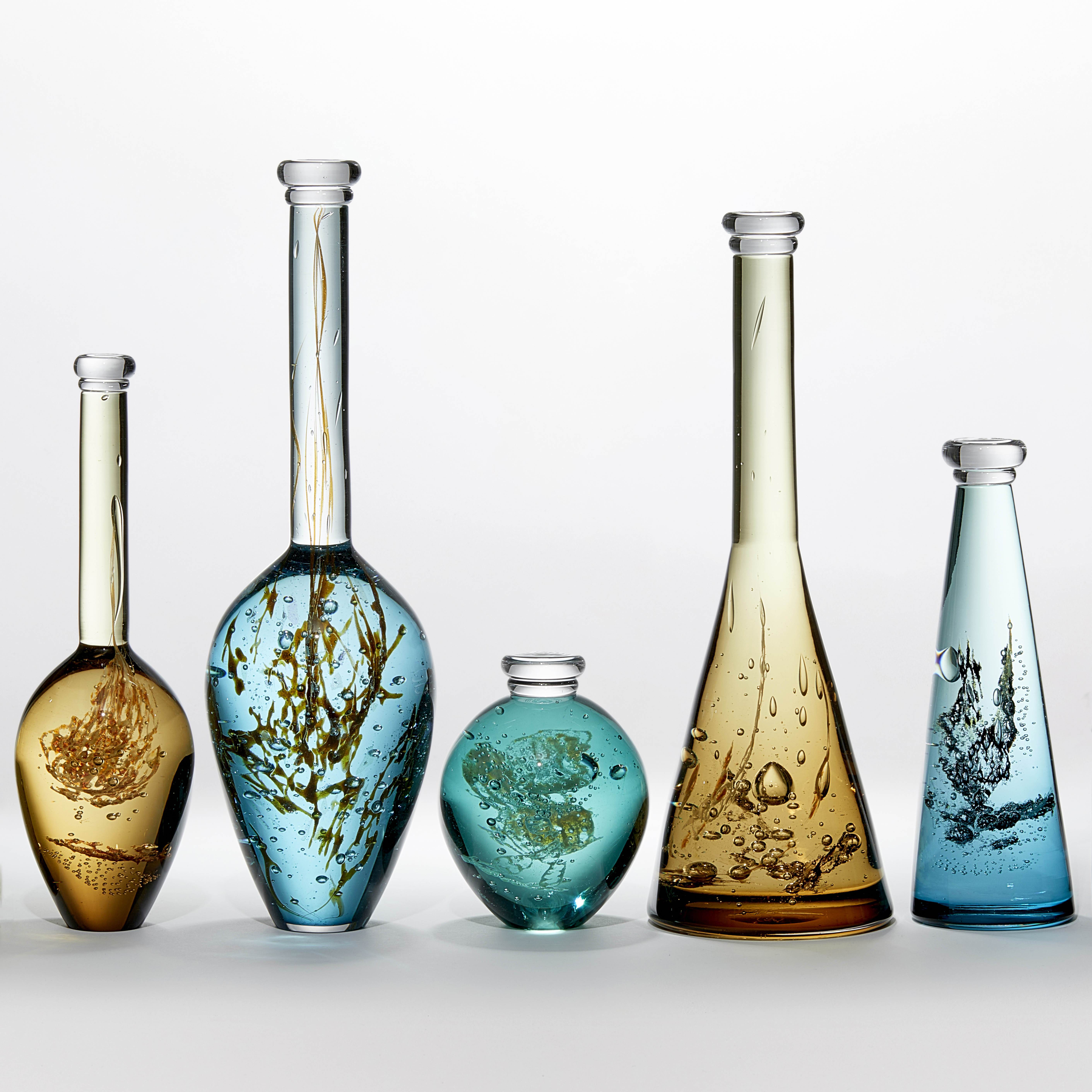 installation of seven solid transparent glass bottles in jade green ochre gold and khaki with organic plant life details trapped inside each with a rounded base and long thin neck and stopper hand made from glass