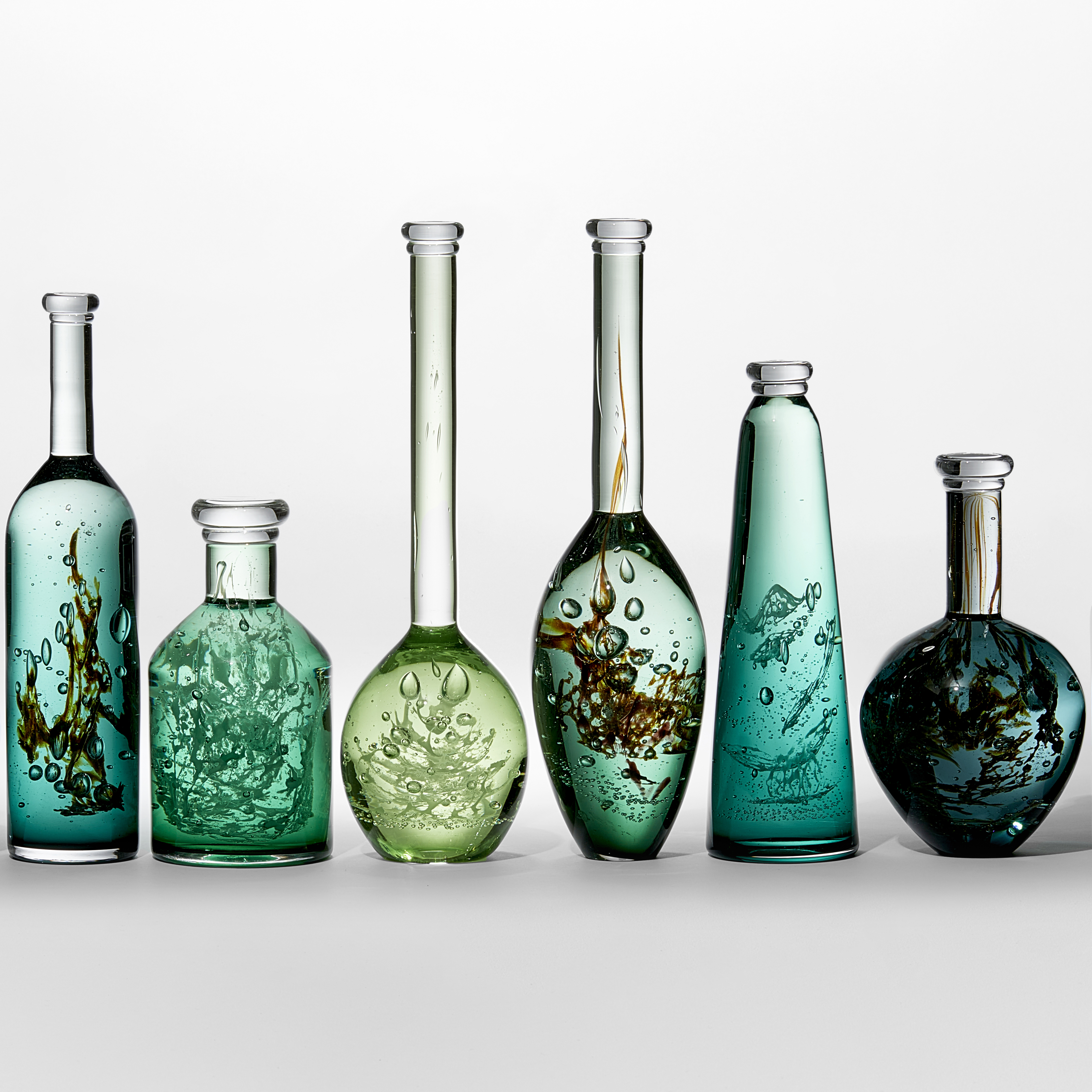 ten unique bottles of different shapes sizes and shades of green with mysterious organic trapped substances and details held within creating a unique installation hand made from glass