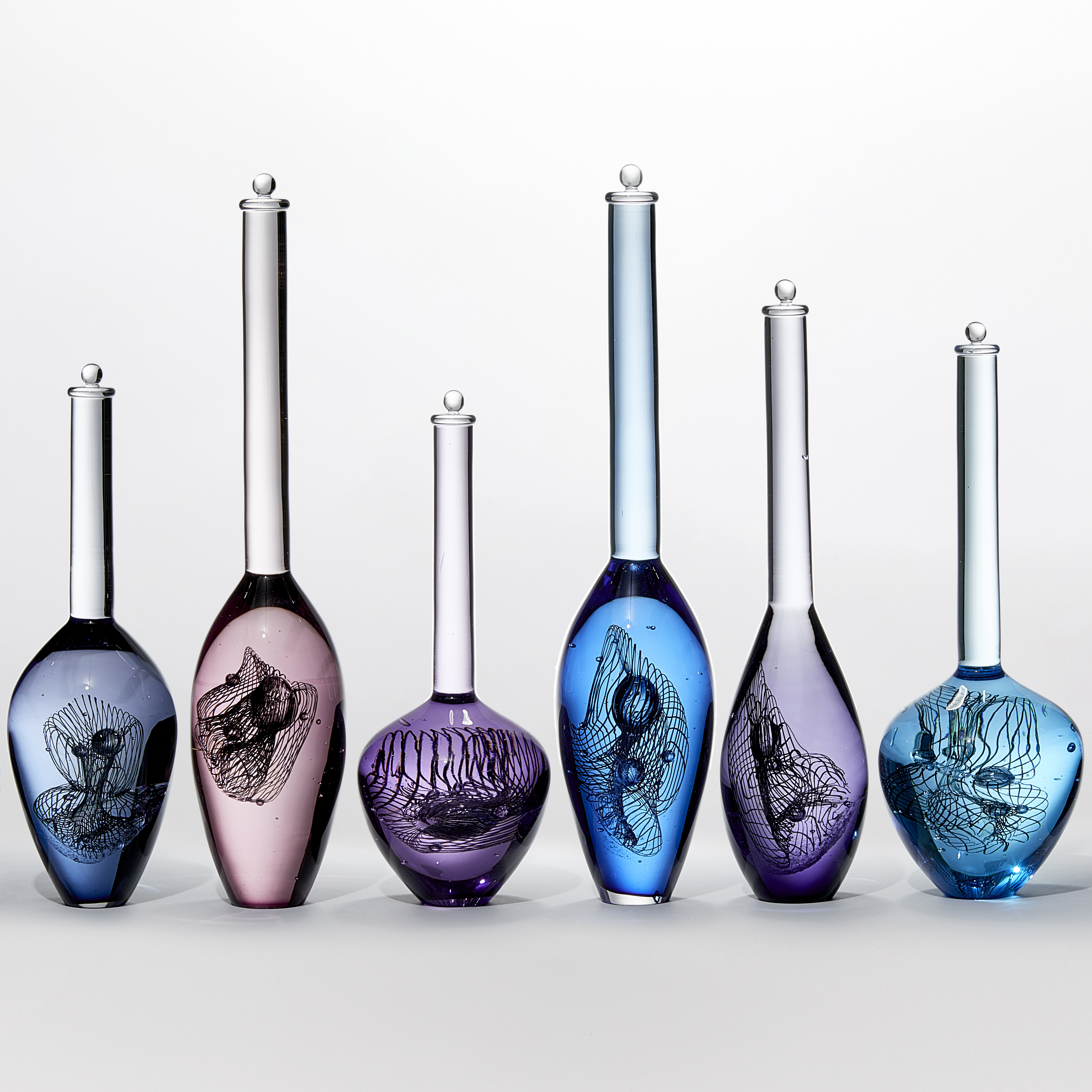 installation of nine solid transparent bottle forms in blue purple and jade each with a long tall neck with stopper capped top and a rounded bottom section containing a double helix hand made from glass 
