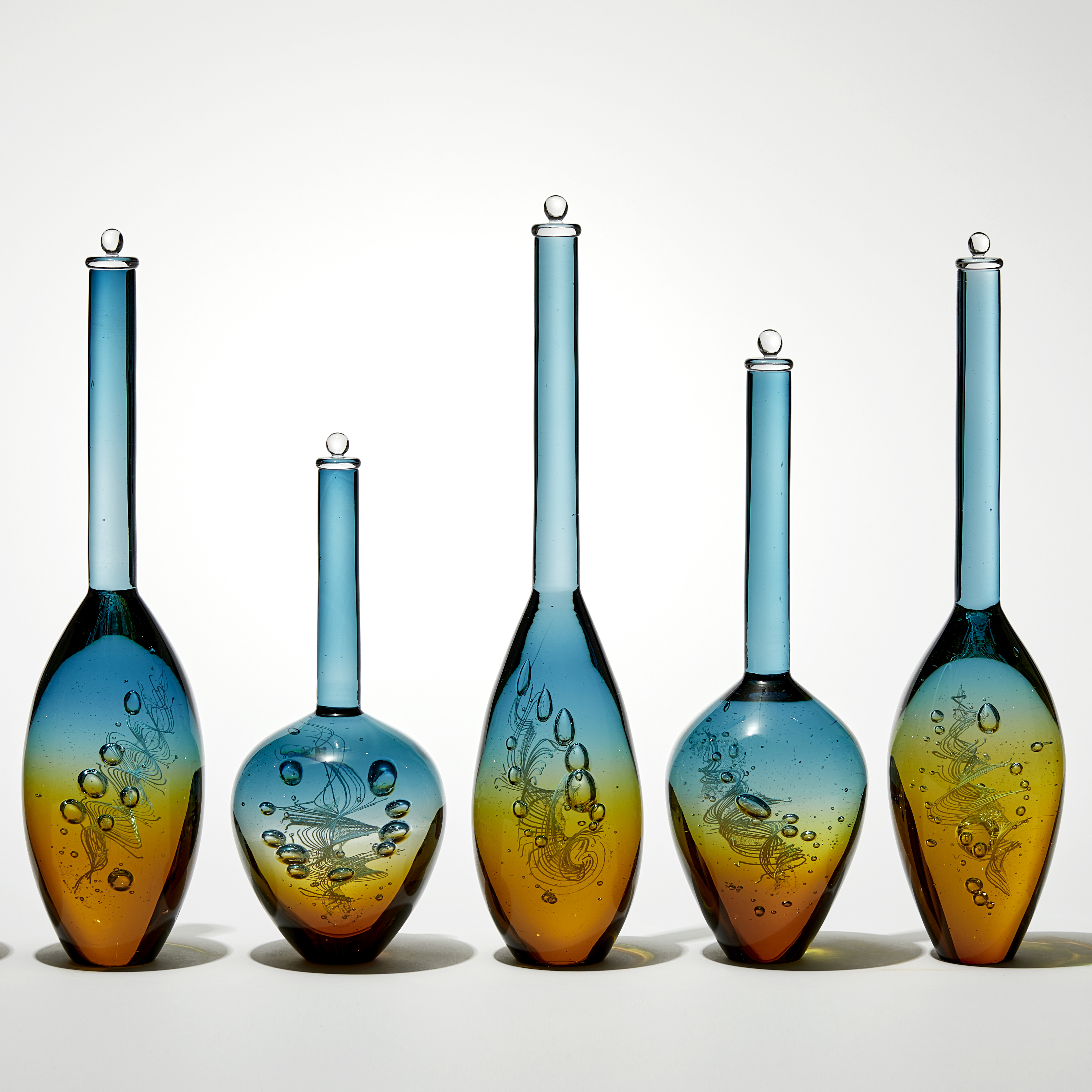 row of seven transparent solid bottles in different bulbous shapes with long thin necks with caps on top with amber bases merging to turquoise tops hand made from glass
