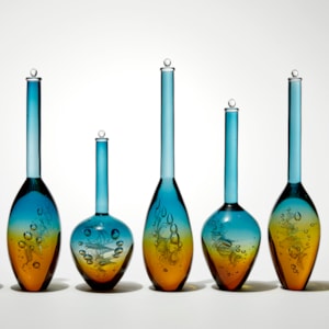 row of seven transparent solid bottles in different bulbous shapes with long thin necks with caps on top with amber bases merging to turquoise tops hand made from glass