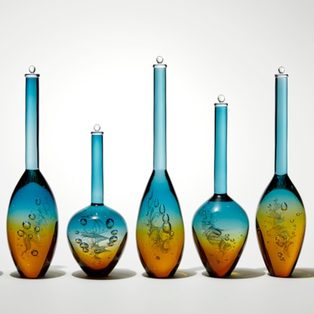 row of seven transparent solid bottles in different bulbous shapes with long thin necks with caps on top with amber bases merging to turquoise tops hand made from glass