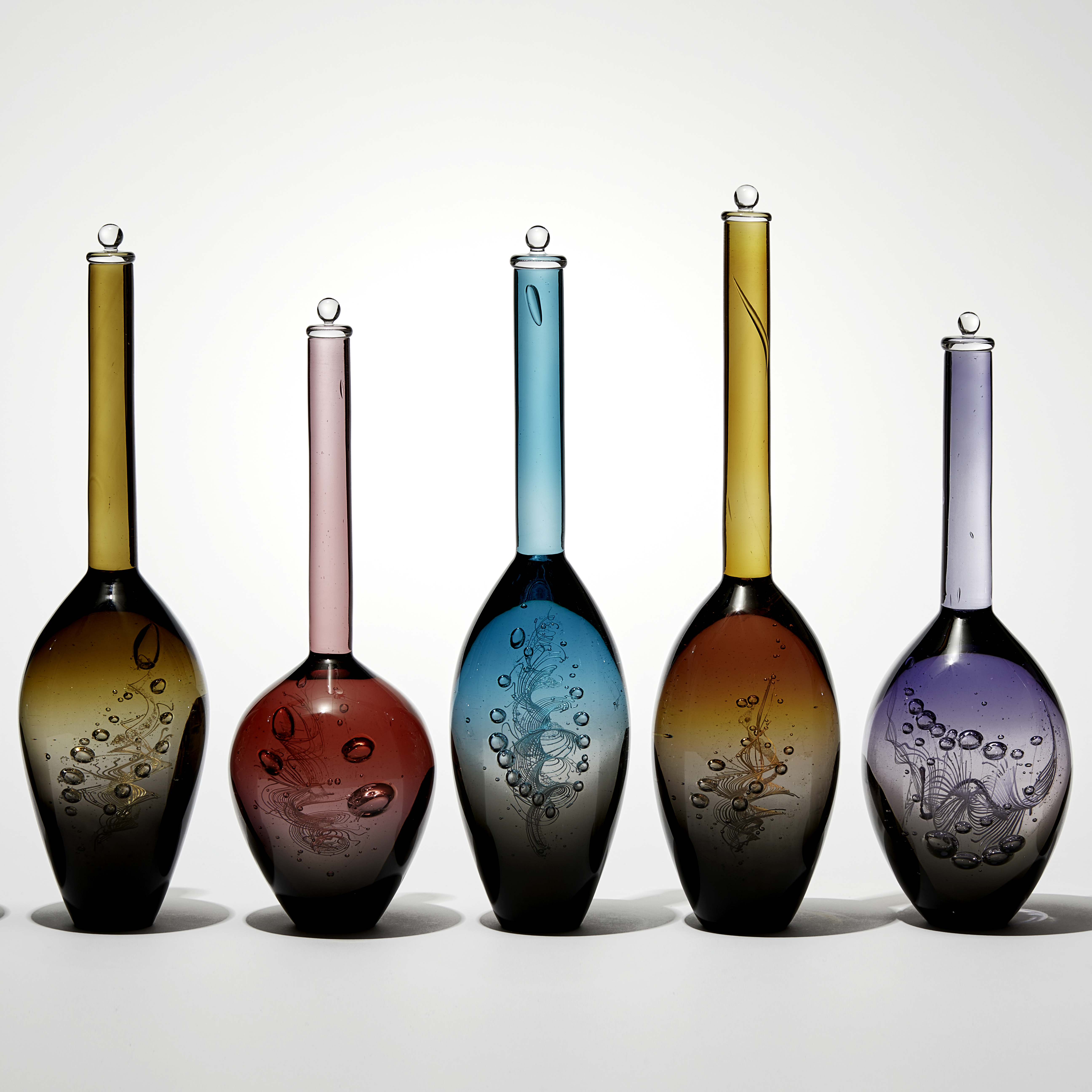 installation of seven transparent solid bottles with interior helix detail each with a grey base merging to a different upper colour all differing bulbous shapes with long thin necks hand made from glass