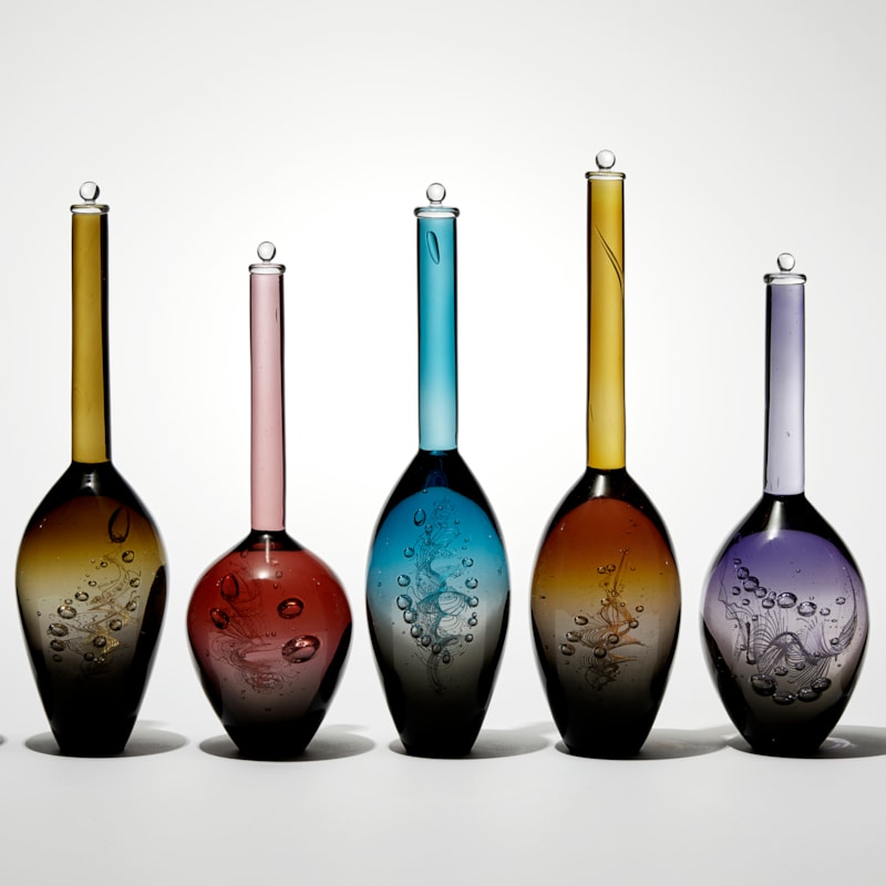 installation of seven transparent solid bottles with interior helix detail each with a grey base merging to a different upper colour all differing bulbous shapes with long thin necks hand made from glass