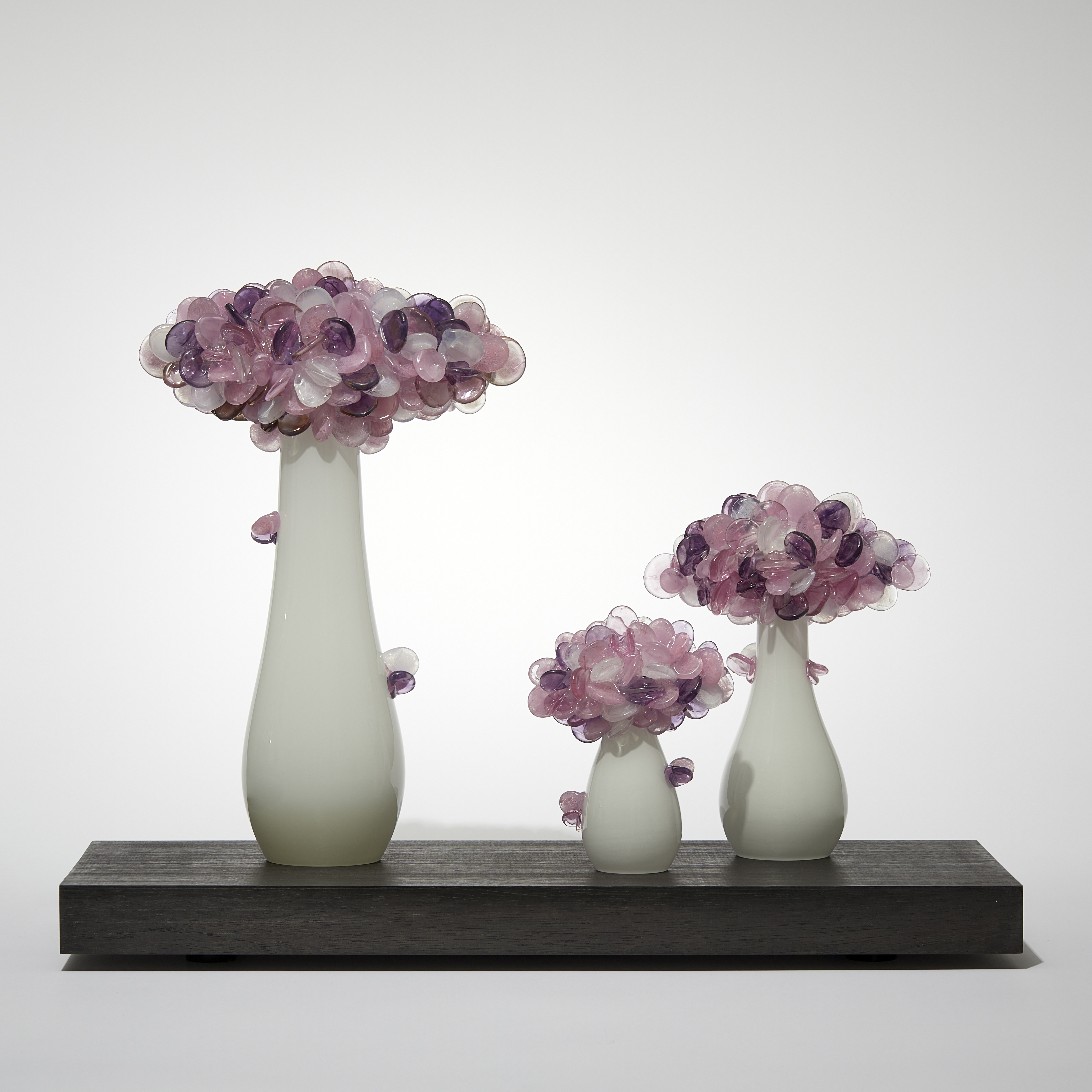 a trio of abstract trees each with an sleek alabaster coloured trunk topped with a multitude of sugary candy cane pink coloured lolliop leaves hand made from glass and presented on a rectangular wooden base