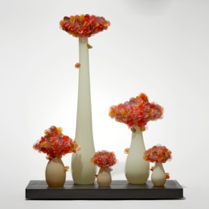 five abstract trees of differing heights with cream sleek trunks topped with clustered orange and amber lollipop shaped leaves hand made from glass and presented like bonsai on a rectangular wood base