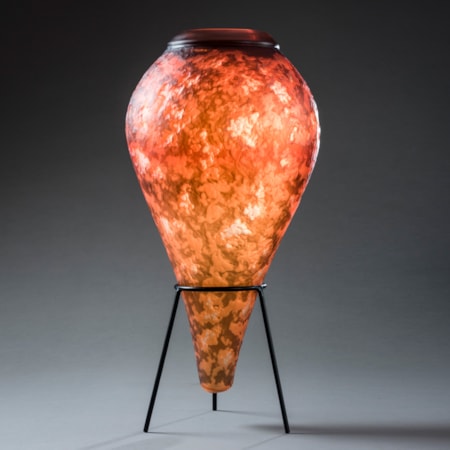 inverted teardrop shaped vessel with lava like textured mottled surface in fiery orange brown and yellow with top opening with a thick black ring hand made from glass sat upon a black steel tripod stand