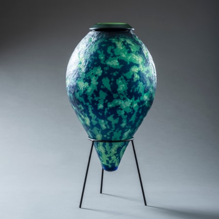 mottled organic textured rich dark blue and jade green inverted teardrop shaped vessel with top opening and think black rim hand made from glass and sat upon a black steel tripod stand