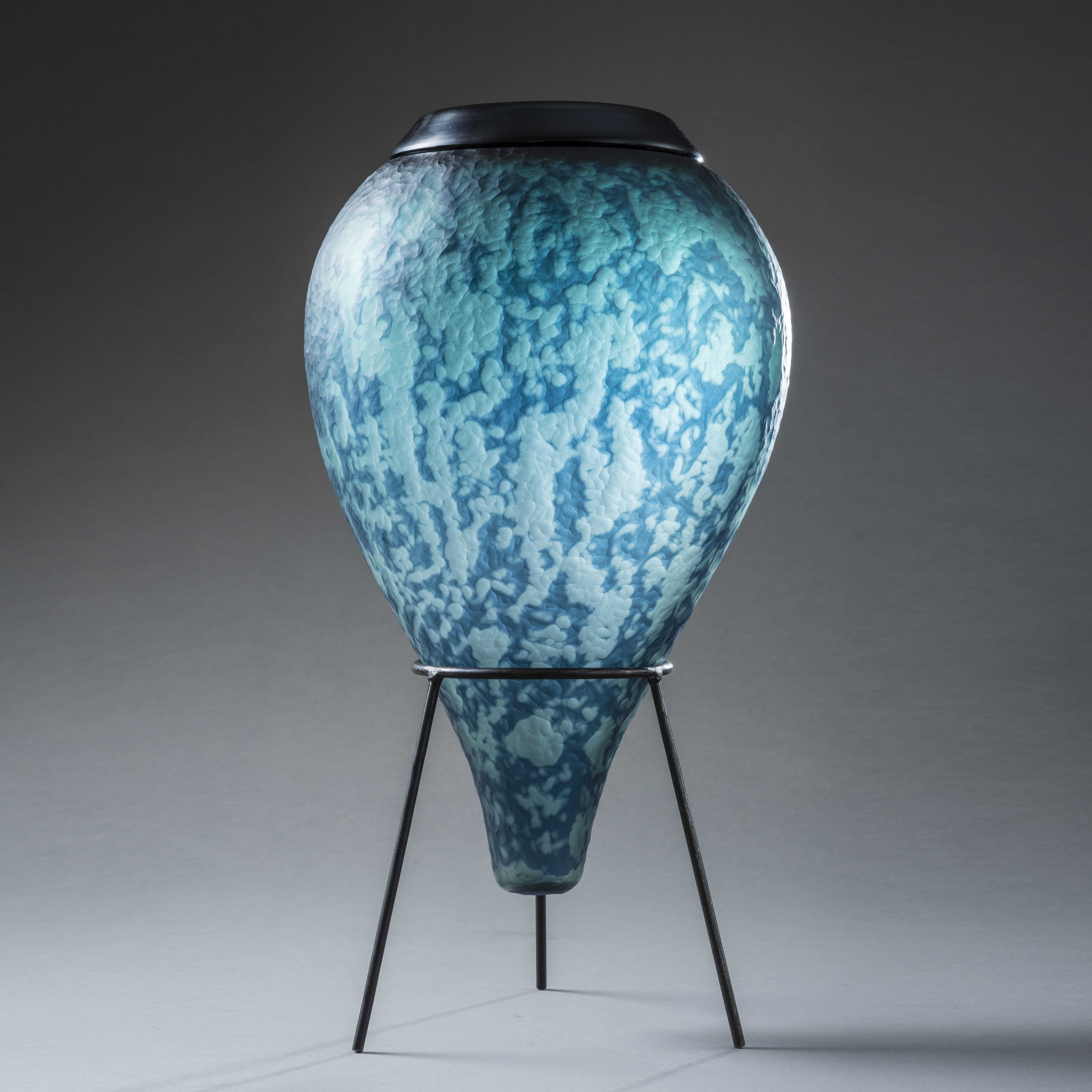 inverted teardrop vessel in mixed aquas and blues with soft mottled organic surface texture and pattern with top opening with smooth black rim hand made from glass with black steel tripod stand
