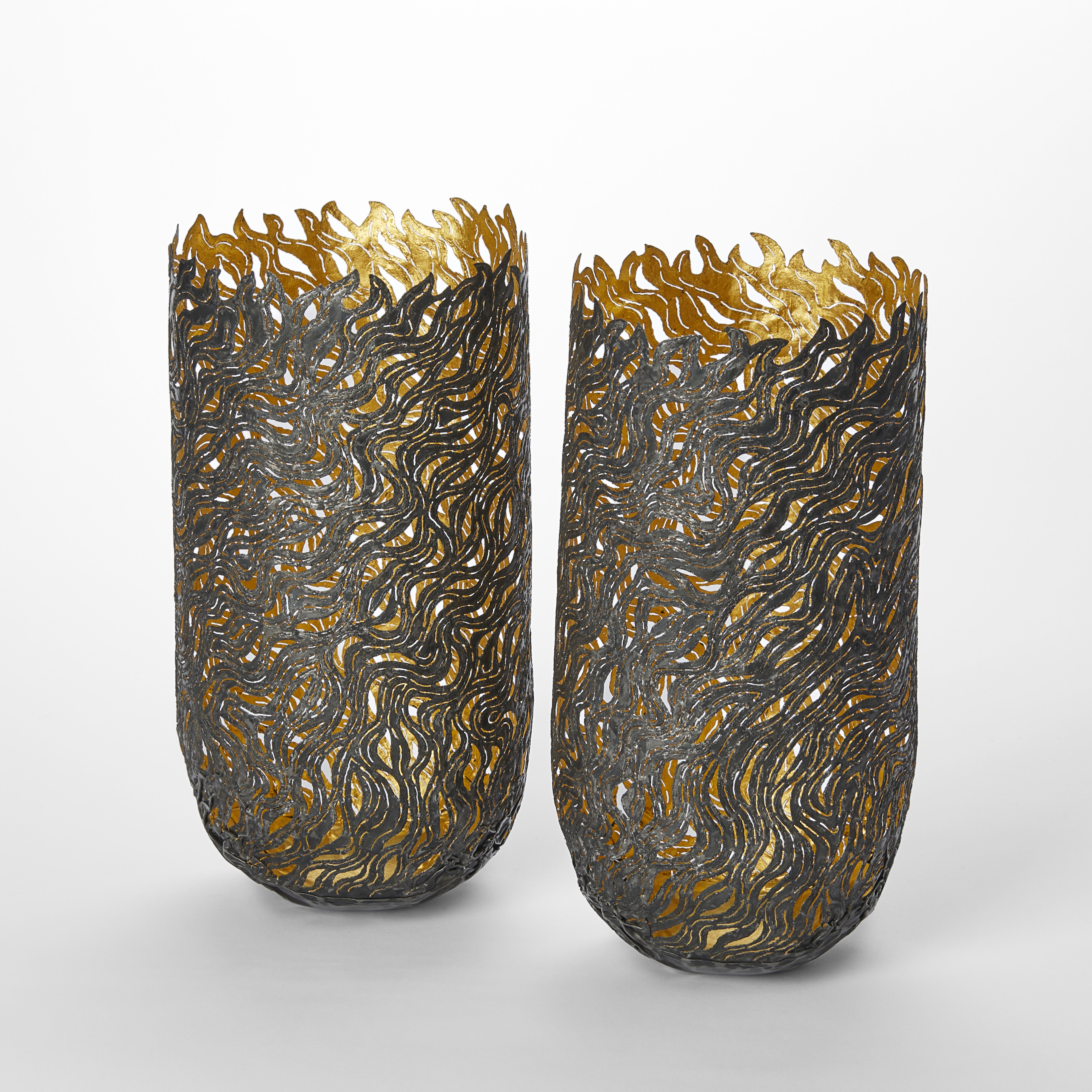 tall darkened steel vessels with rounded bases and jagged flamed shaped top edges with arced rounded curling patterns on the surface and cut out sections with a gold reflective interior surface