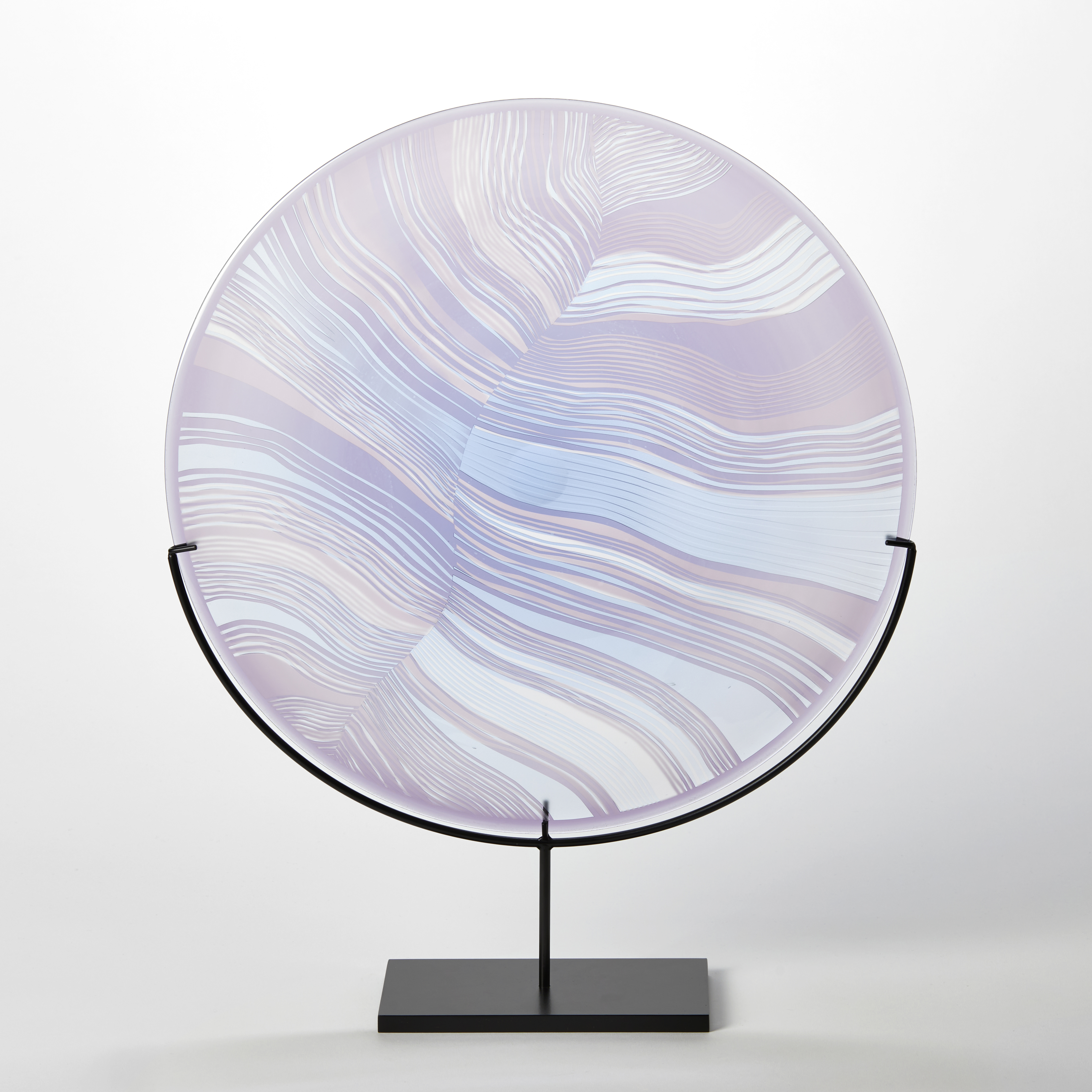 round translucent rondel in soft lilac and aqua with abstract cut lines on the surface similar to a birds feather handblown from glass presented on a matt black stand