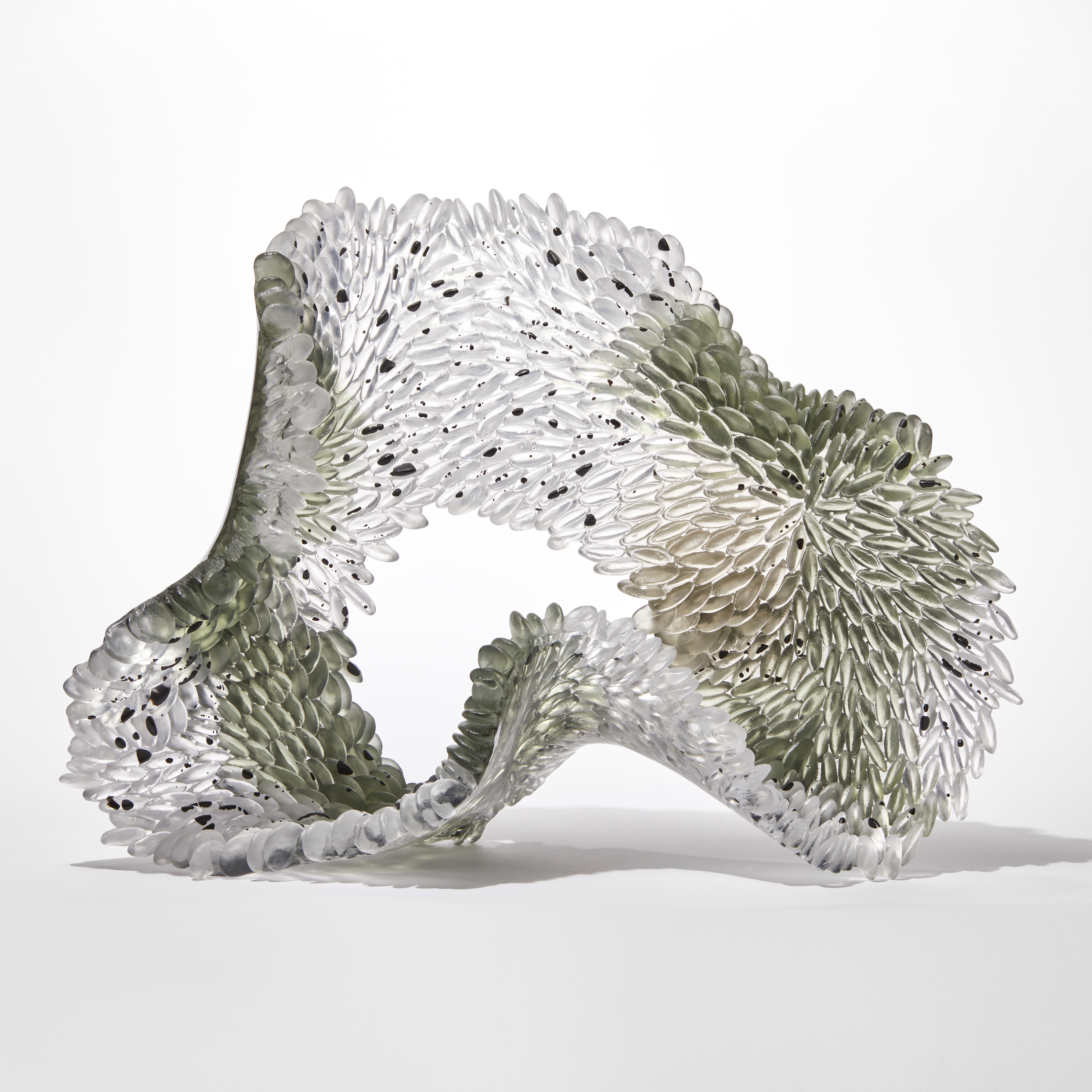 clear textured organically looped standing glass sculpture with one side scaled the other smooth with areas in clear soft moss green and bronze with speckles and dots of opaque black