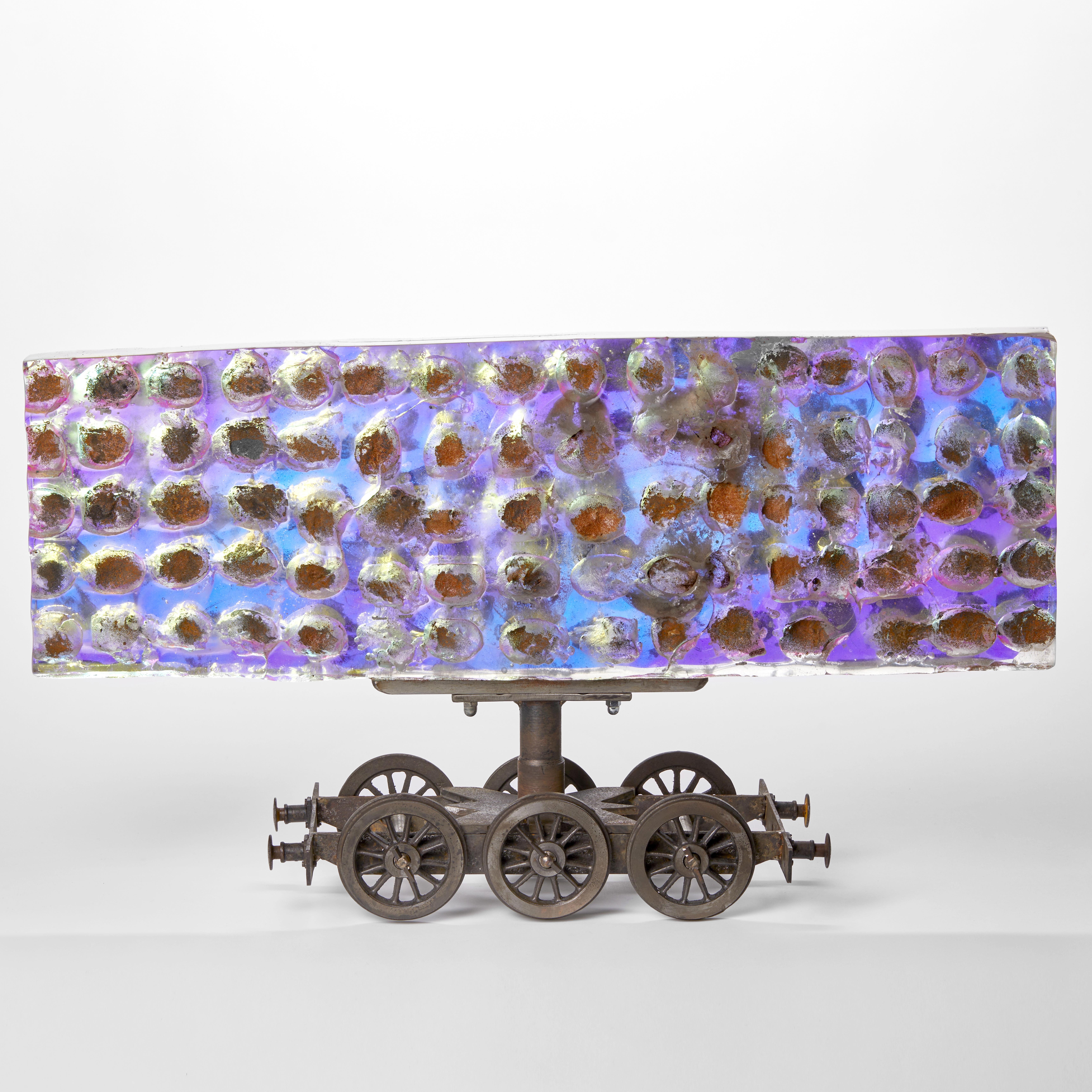 steel six wheel train chassis with a large rectangular bill board shaped sheet of thick cast glass above with sandwiched filter in the centre creating a shimmering and emitting a spectrum of colours in blue greens lilacs and purples with a rough dimpled surface with rusted wells