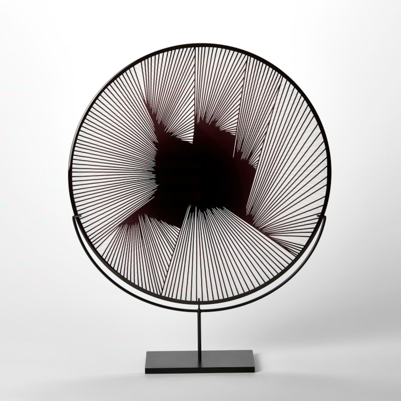 round clear and black hand blown glass decorative plate with abstract linear geometric etched lined pattern on the surface presented on a black steel stand
