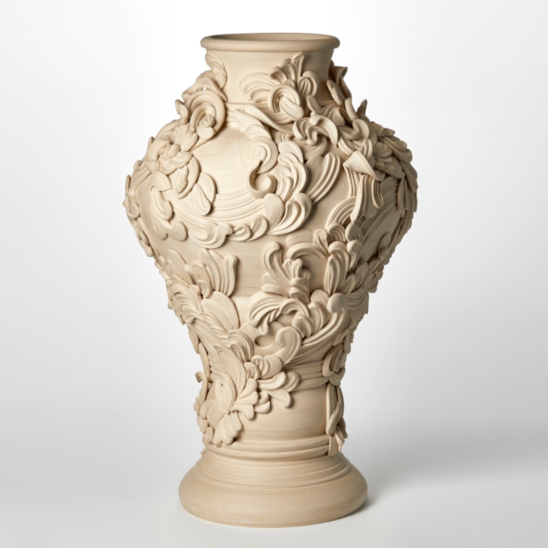 tall classical sandstone coloured vase with narrowing base bulging upper middle and top narrower round opening covered with architectural rococo swirls and flourishes hand made from white st thomas clay