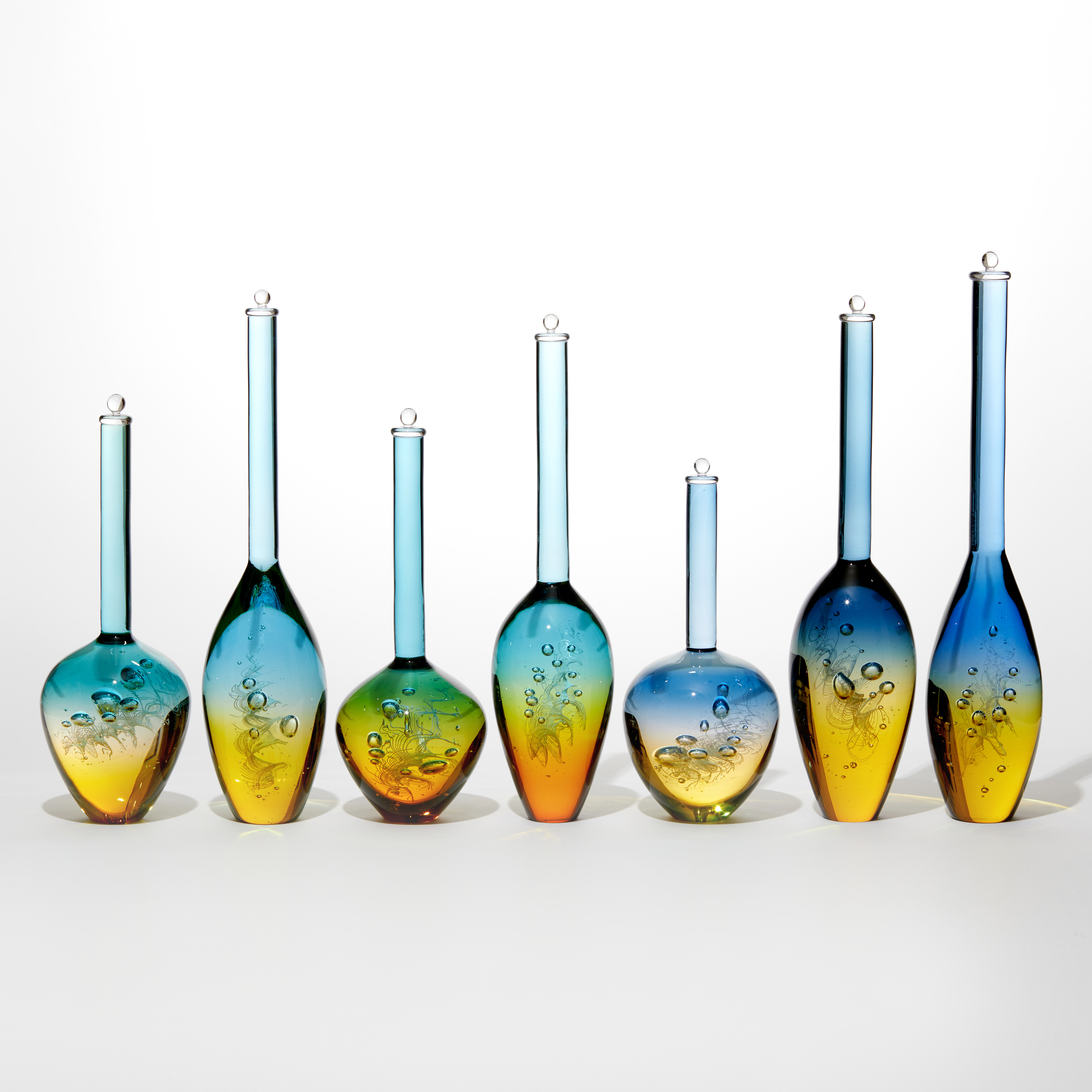set of 7 different shaped solid transparent bottles with trapped bubbles inside each with a golden yellow bottom half merging to bright blue uppers all with long necks and stoppers on the top