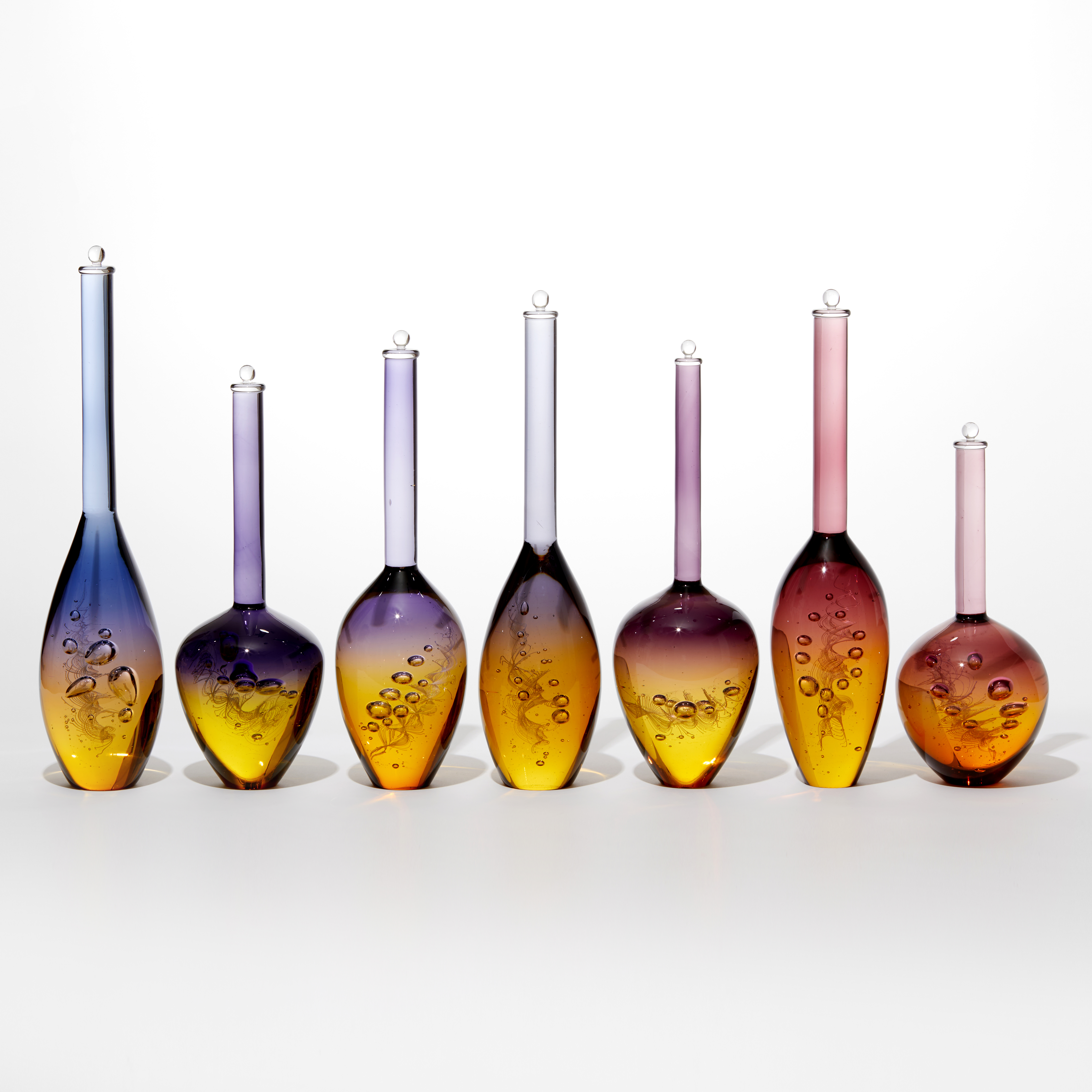series of seven solid glass bottles with bubbles trapped inside in a helix spiral all slightly different in size and height with gold bottoms merging to purple tops with long necks and stoppers on the top