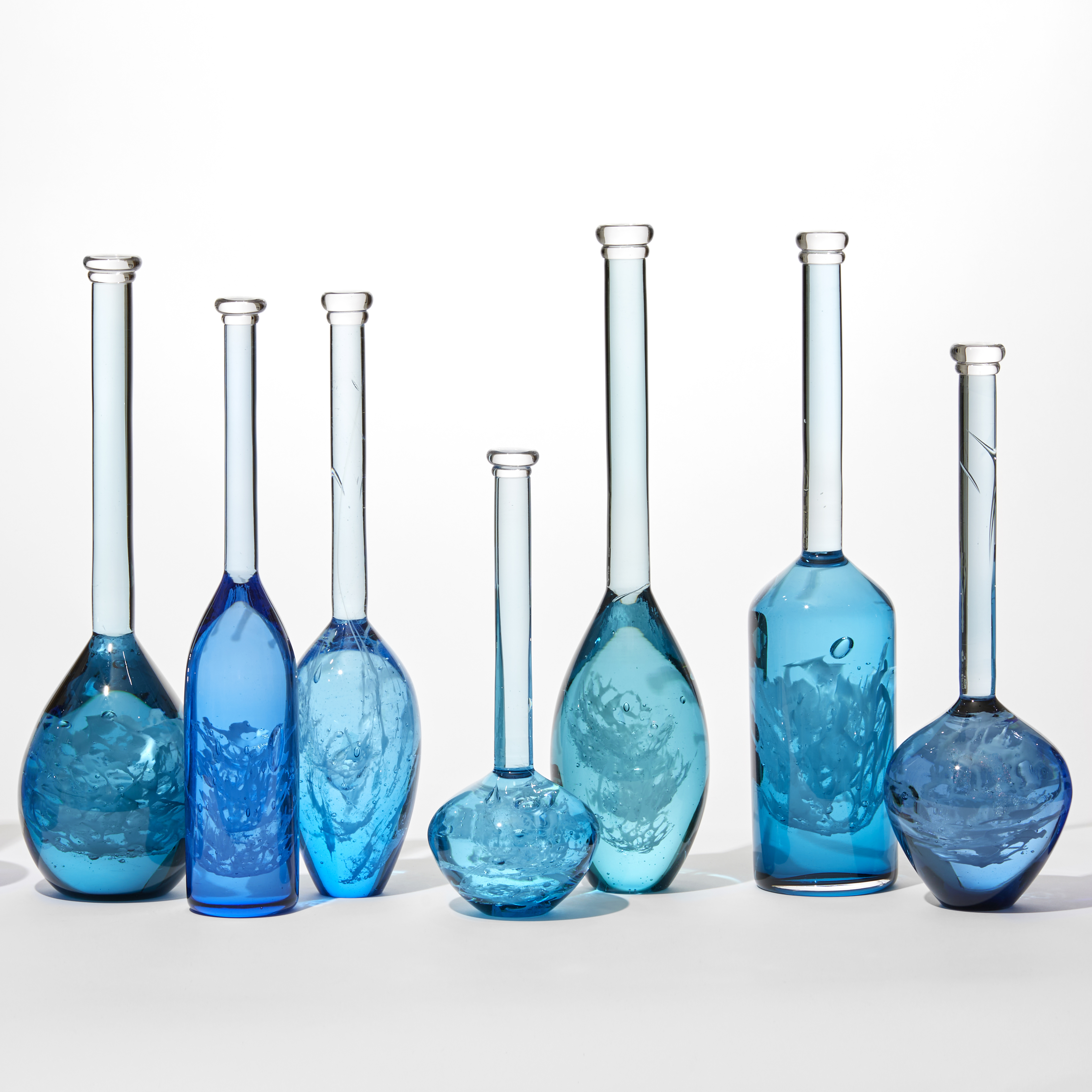 installation of 10 solid bottles in varying hues of jade royal blue aquamarine and turquoise with the suggestion of swirling mysterious contents held within each one all having long thin necks and caps on the top hand blown from glass