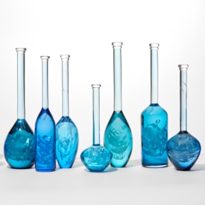 installation of 10 solid bottles in varying hues of jade royal blue aquamarine and turquoise with the suggestion of swirling mysterious contents held within each one all having long thin necks and caps on the top hand blown from glass