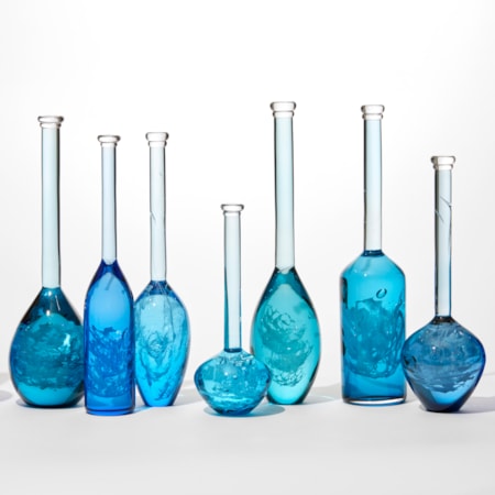 installation of 10 solid bottles in varying hues of jade royal blue aquamarine and turquoise with the suggestion of swirling mysterious contents held within each one all having long thin necks and caps on the top hand blown from glass