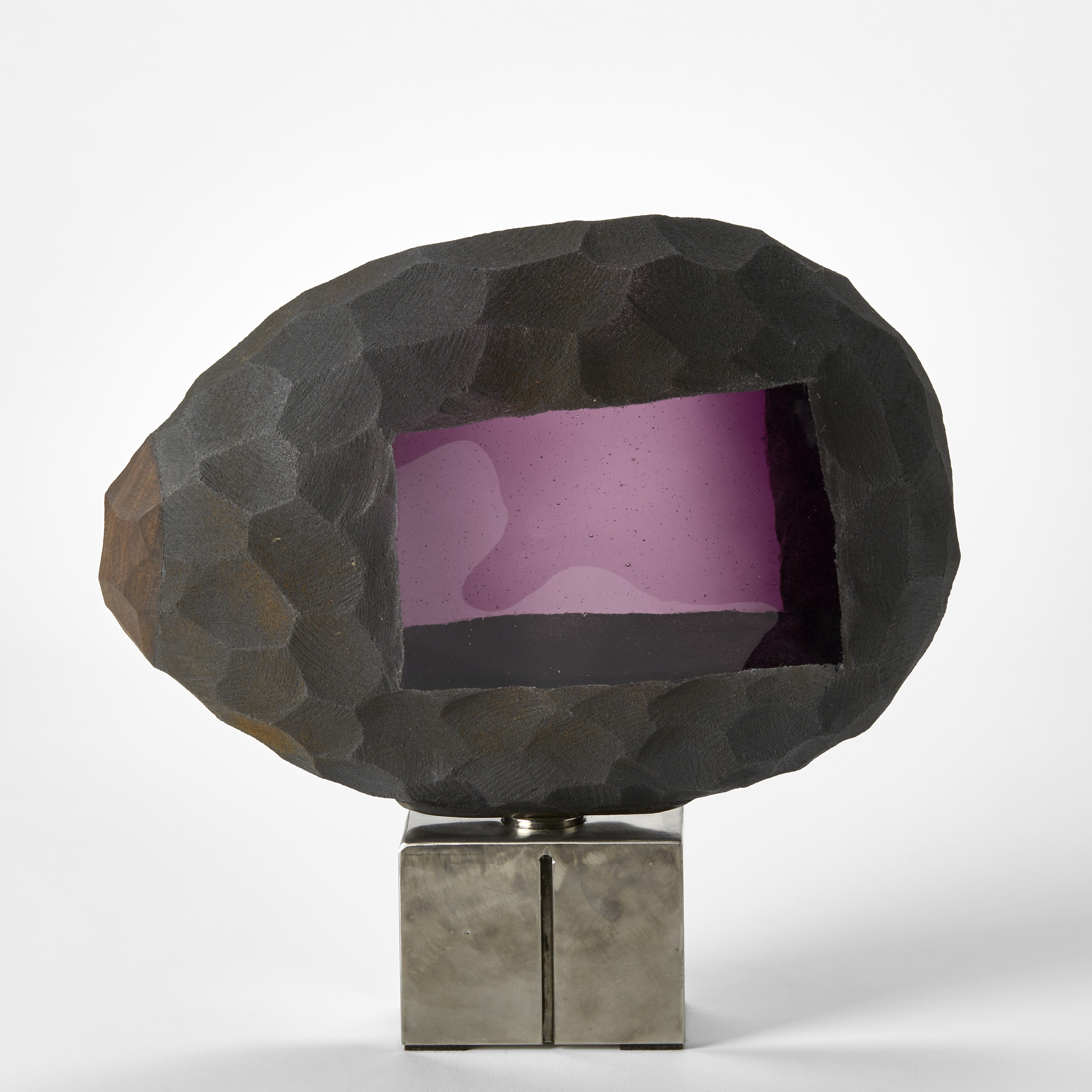 oval rough bubble of glass with rough cut exterior with dark metallic finish and deep incised scallops with central wonky rectangular purple window revealing the interior with a solid steel cubed base with a magnet holding the two in place stacked on on top of the other hand made from glass