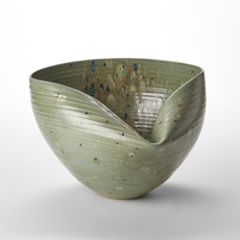 round hand thrown bowl with one folded in side creating a neat inward tuck with slight horizontal ridges on the surface in a soft sage green colour with dark blue smudgy dots hand thrown from white saint thomas clay