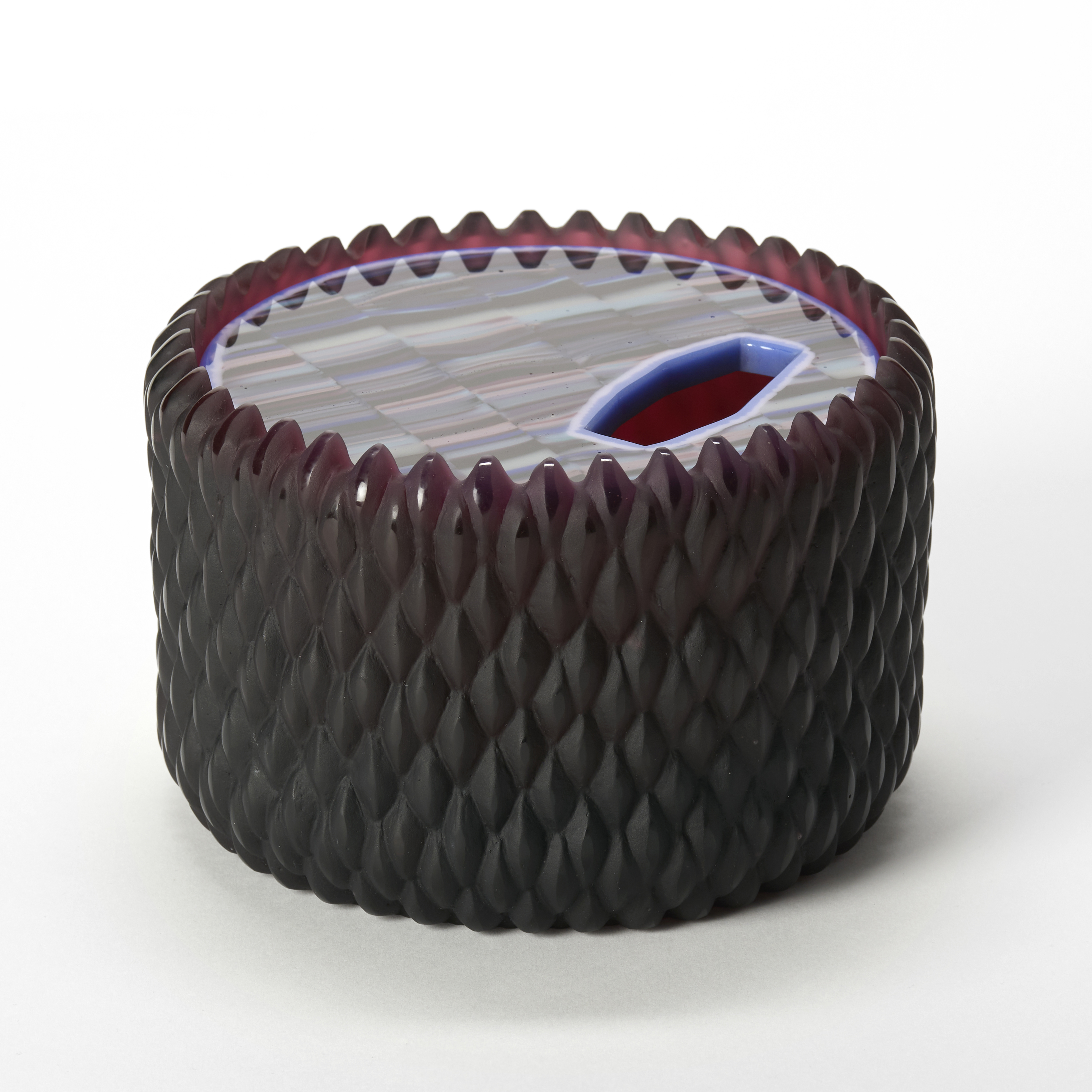 round low lidded sculptural jar with a matt base in opaque aubergine purple with a relief repeat diamond pattern and pointed top edge with a round  insert lid with shiny top and strata pattern captured within in jade blue and purple hand made from glass