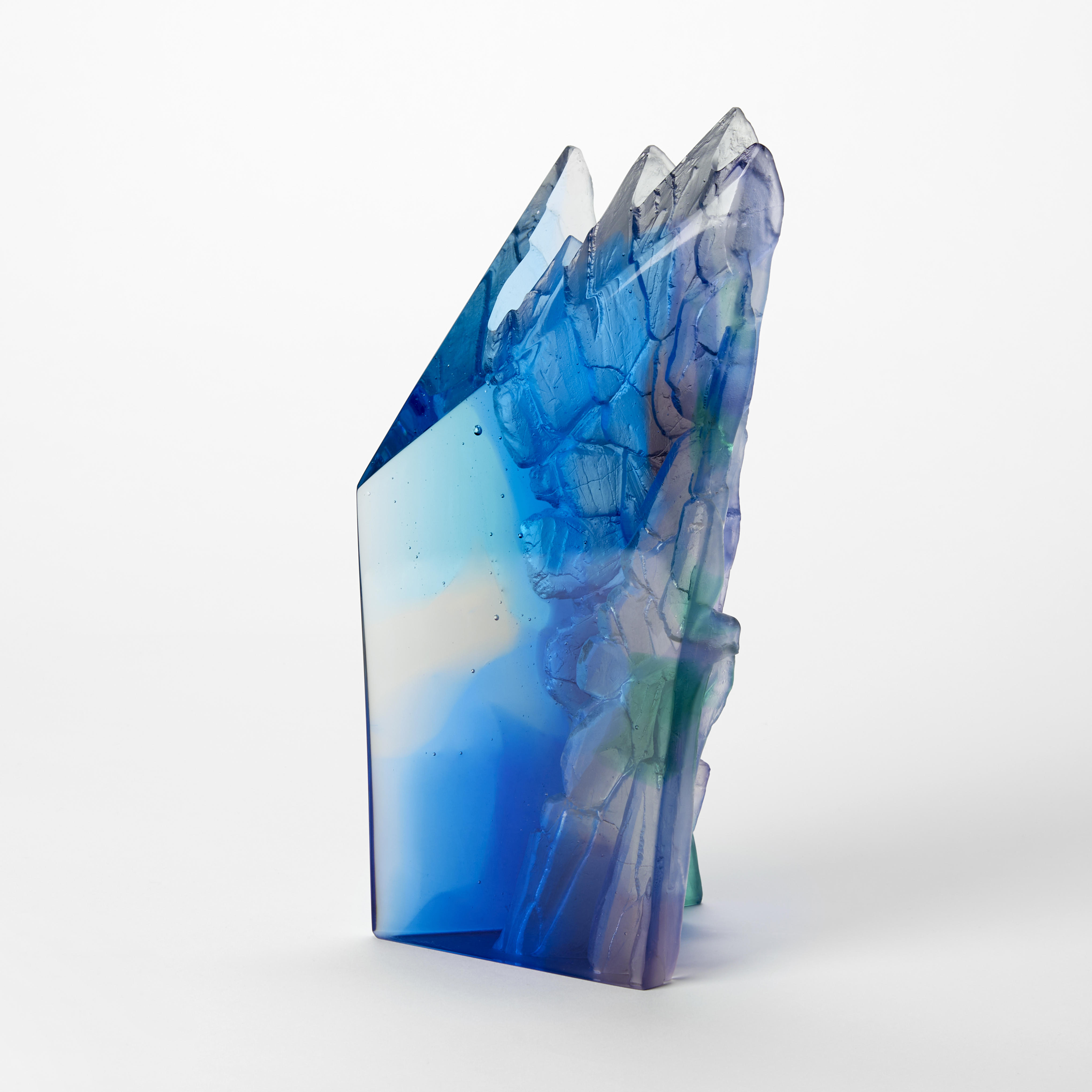 solid transparent standing angular sculpture resembling a cliff with two flat shiny sides and two recessed sides with a craggy organic texture which can be seen through the mass hand made and cast from blue jade green and clear glass