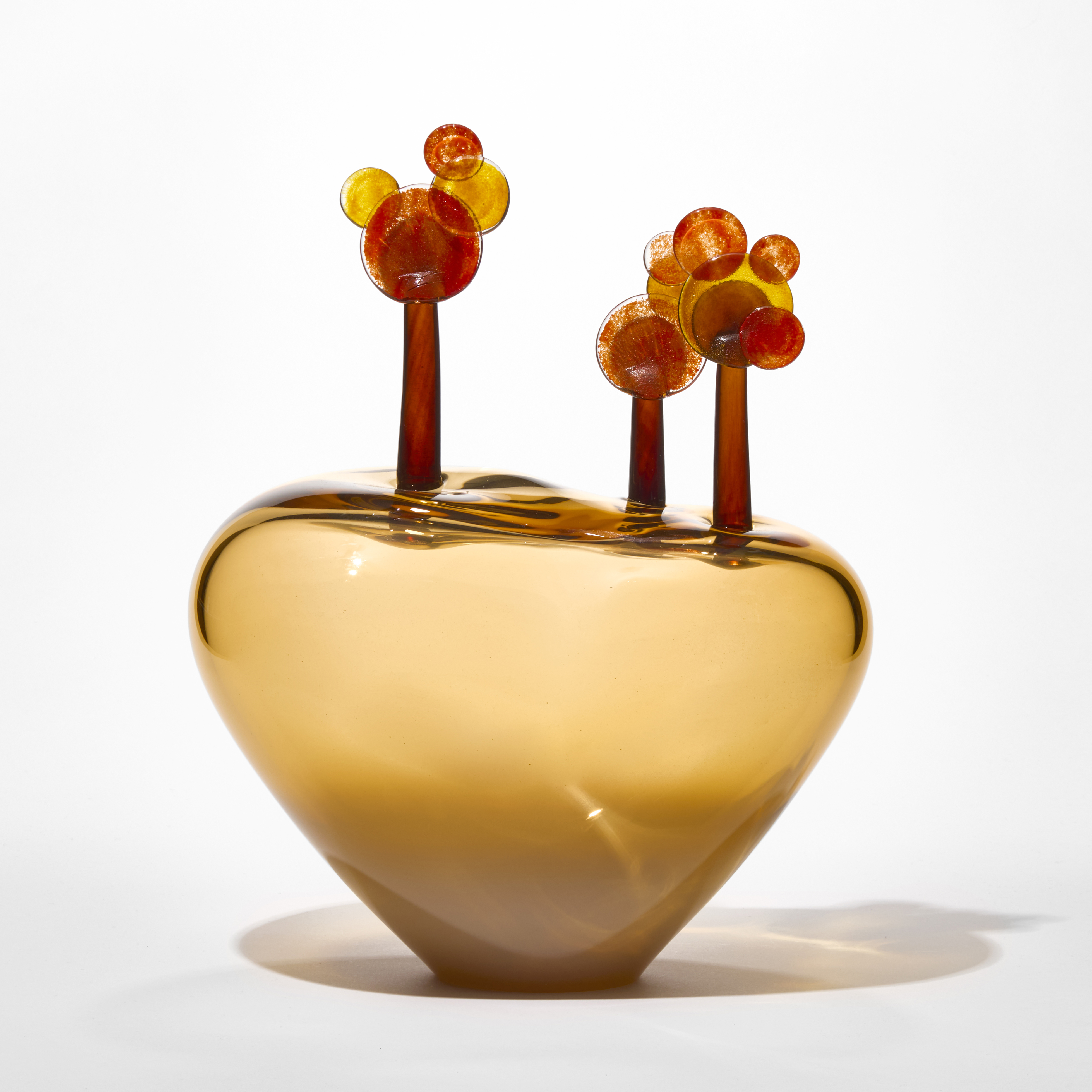 low transparent ochre inverted triangular squat vessel with three red umber and amber trees perched on the top each with a thin trunk and round lollipop leaves hand blown from glass