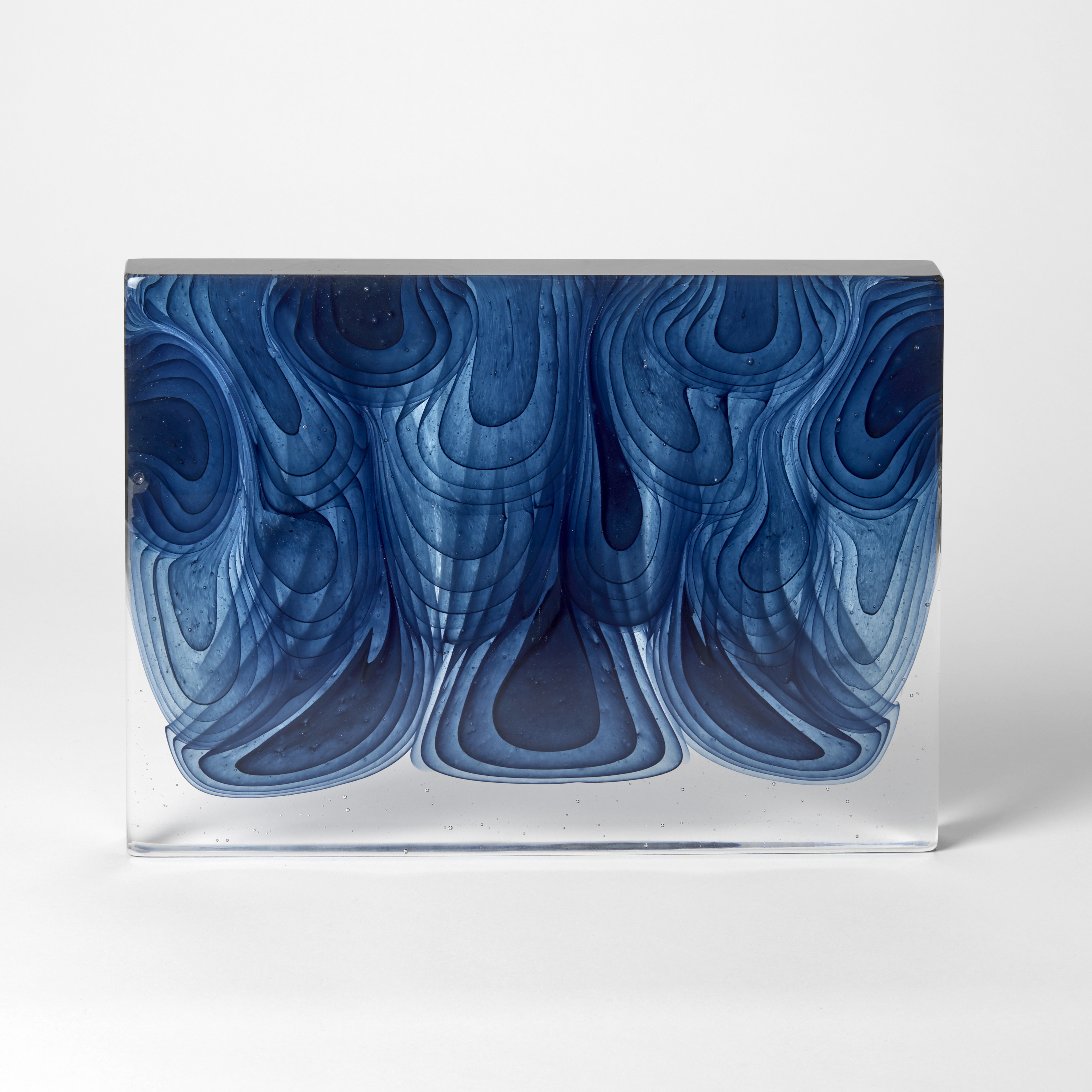 standing thick transparent slab with blue repeating concentric droplet sections in differing sizes pouring from the top towards a clear base hand made from cast glass