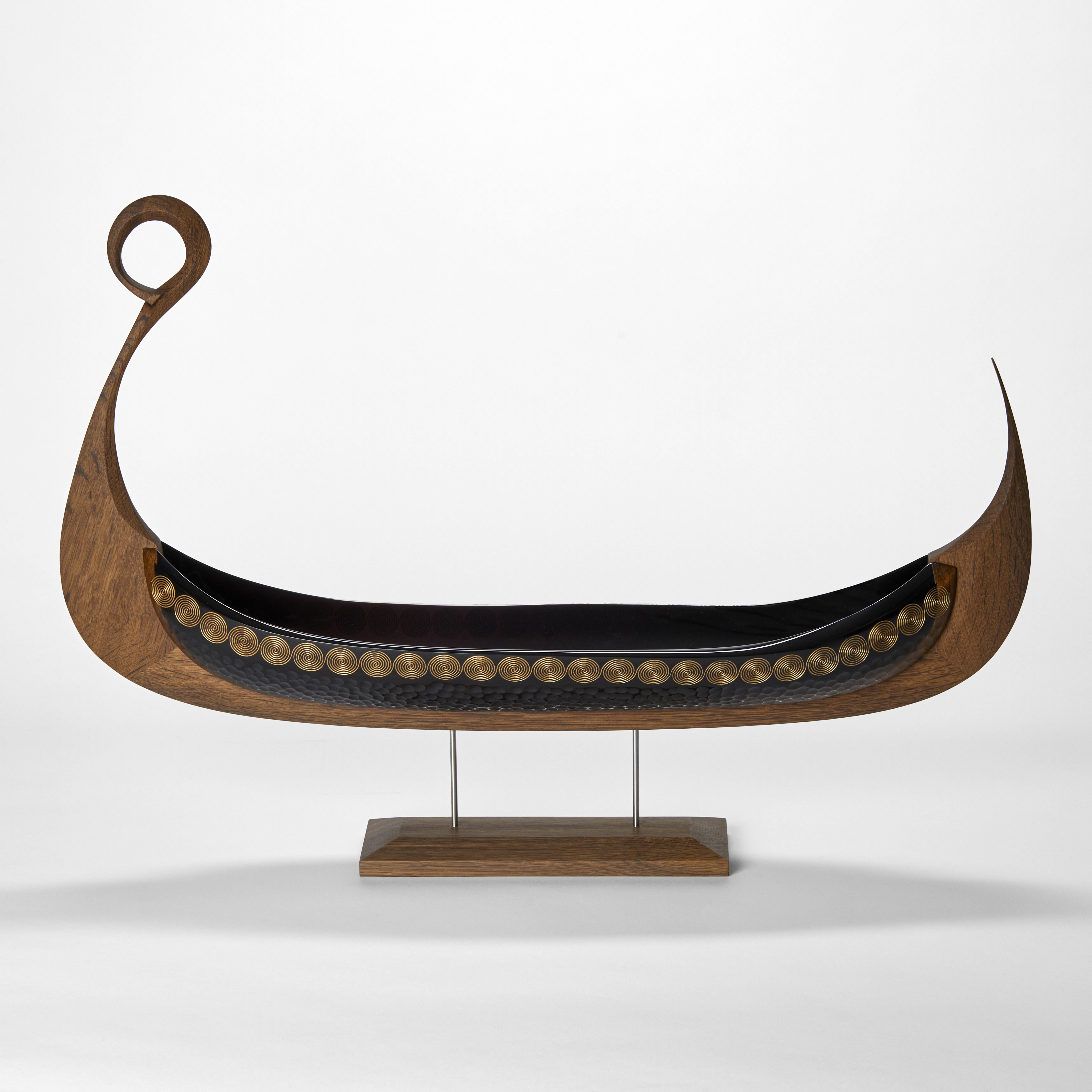 steel blue viking ship artwork with carved oak keel with ring detail at the front with a cut and blown glass hull with decorative patterns engraved on the surface presented perched on a simple stand