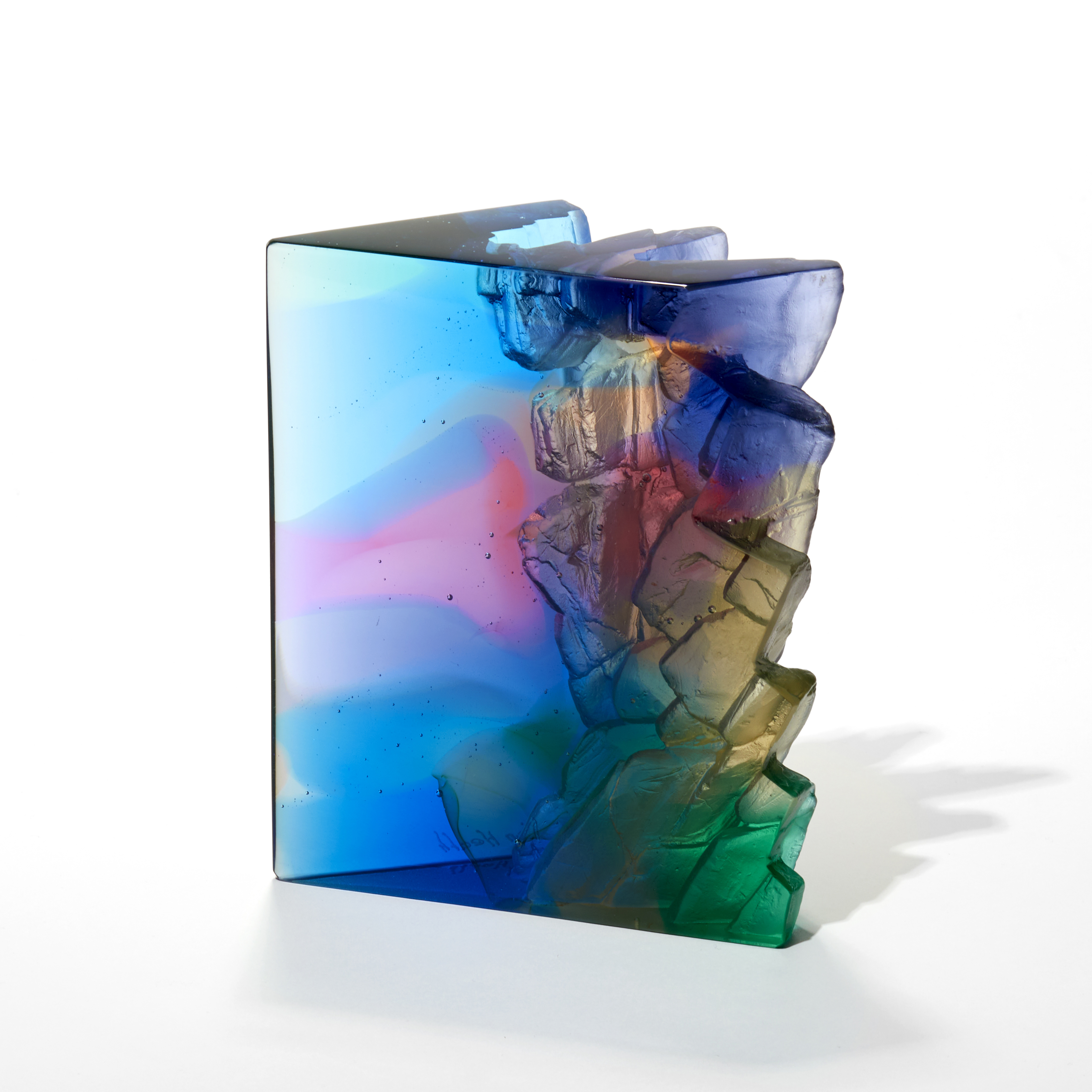 transparent standing form in watery aquas and rich blues with organic merging areas in pink green and taupe with a flat top and two polished vertical sides with one side with a craggy rock like texture hand made from cast glass