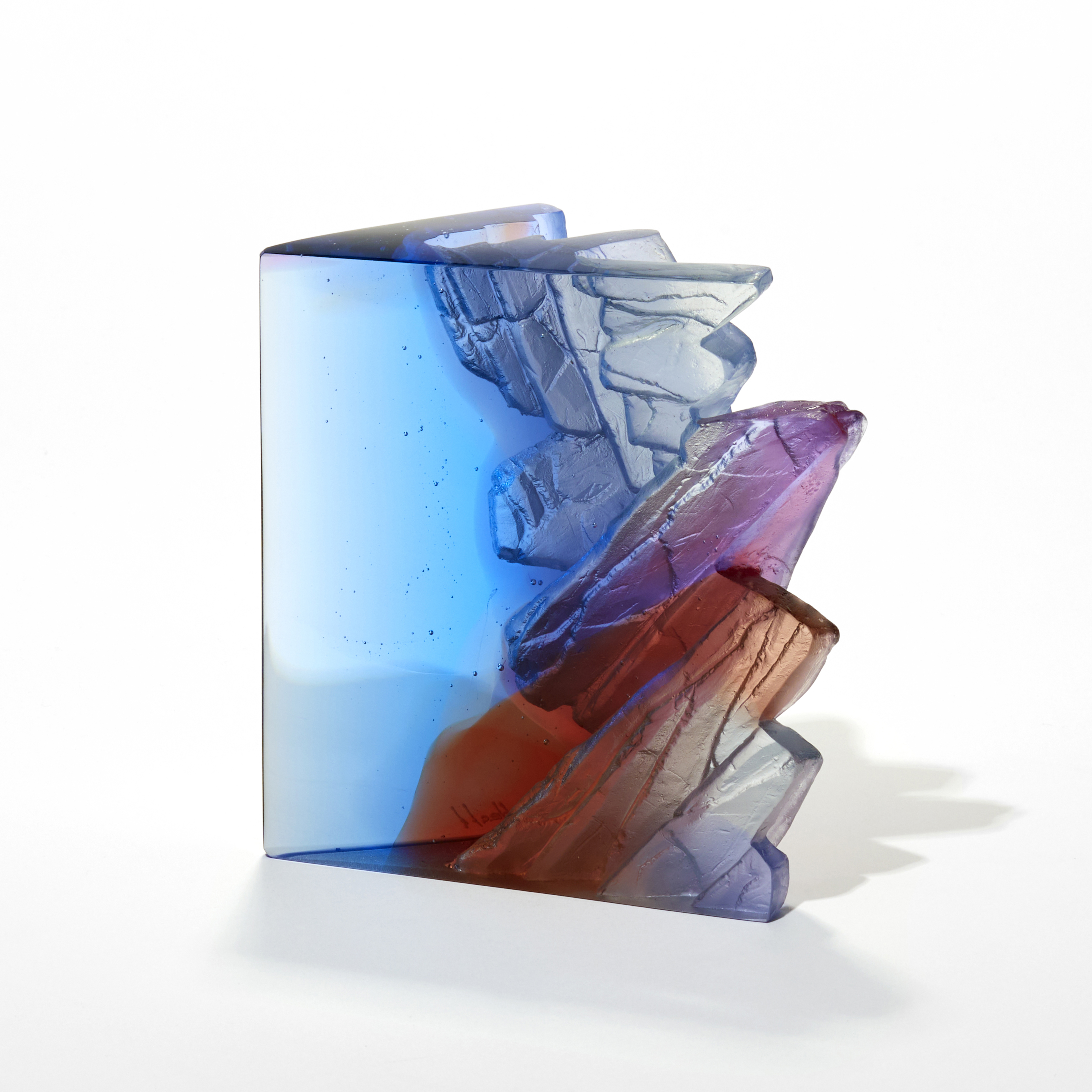 chunky standing transparent sculpture in fading blue rust peachy red and lilac with two vertical glossy sides and top and one side with a textured rock like surface hand made from glass